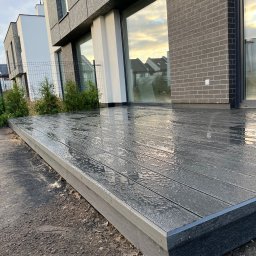 Vk-Bud - Nowoczesny taras z desek kompozytowych w kolorze grafitowym, po deszczu. Widoczne odbicia w mokrej powierzchni, minimalistyczna architektura domu w tle.