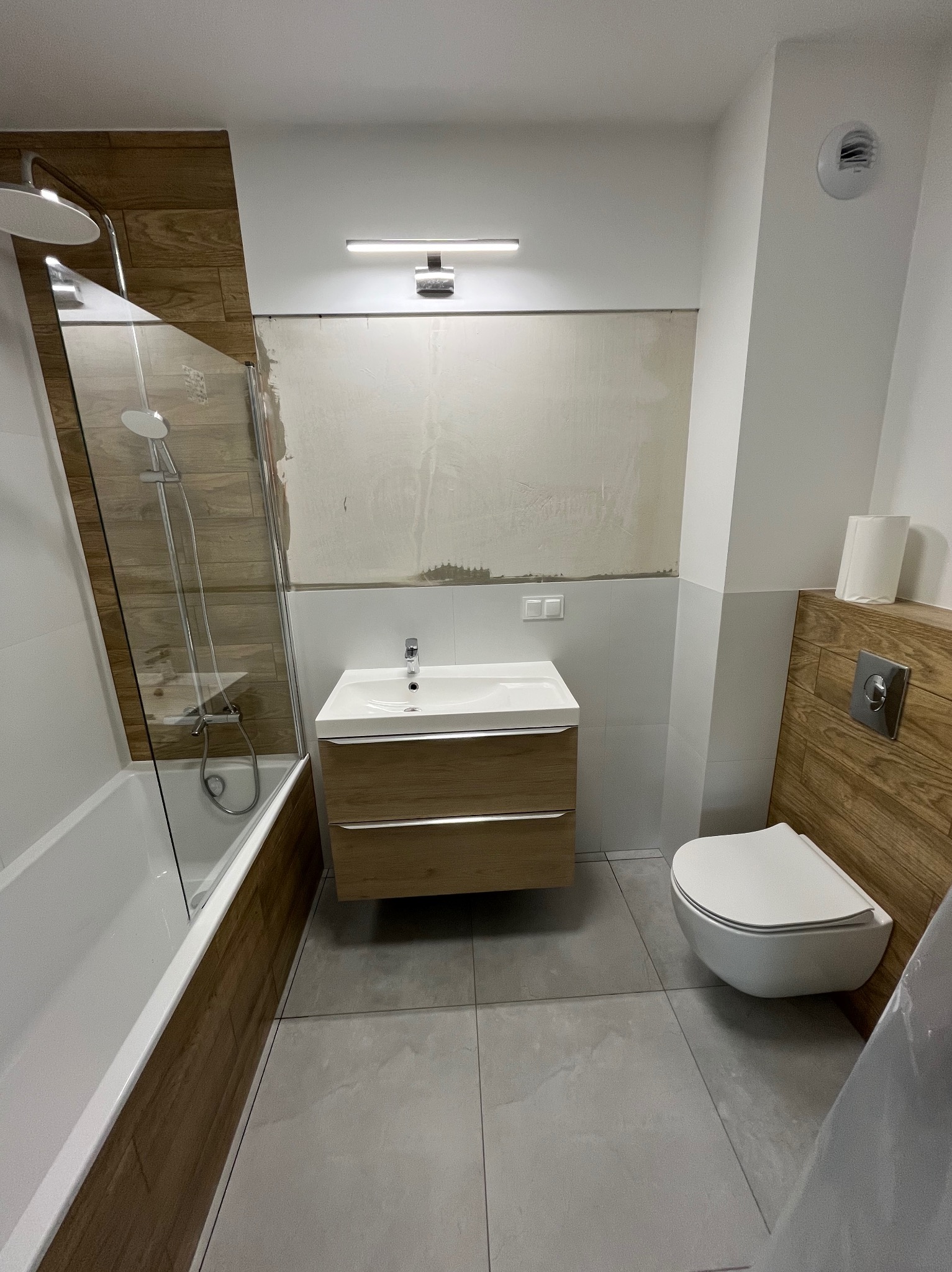 Nowoczesna łazienka w trakcie wykańczania: drewnopodobne płytki, biała armatura (wanna, umywalka, toaleta), szare płytki podłogowe. Częściowo położone płytki na ścianie.