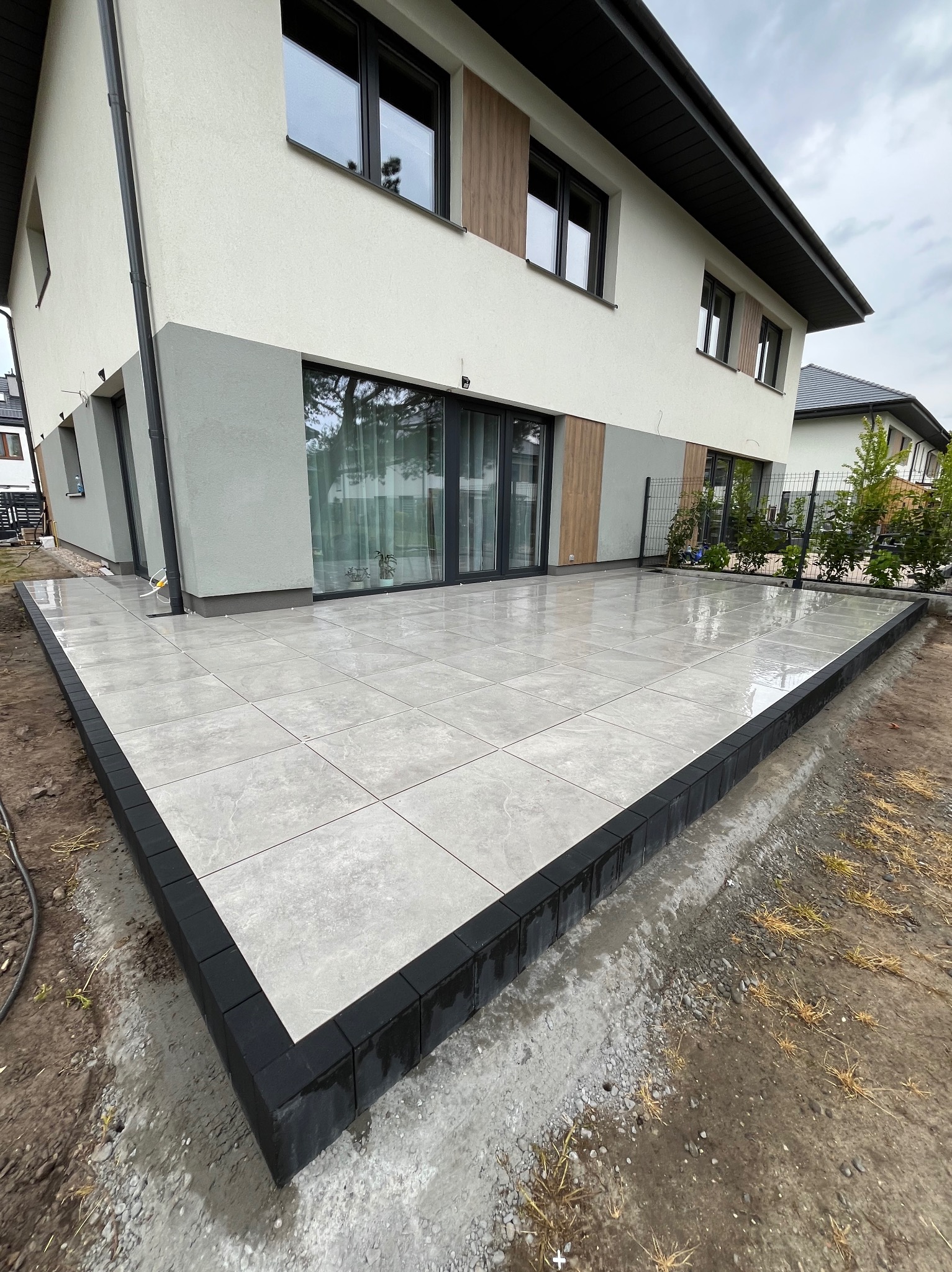 Nowoczesny taras z jasnoszarych płytek, obramowany czarnym obrzeżem, przylegający do budynku z dużymi oknami. Widoczne detale wykończenia i świeżo ułożona nawierzchnia.