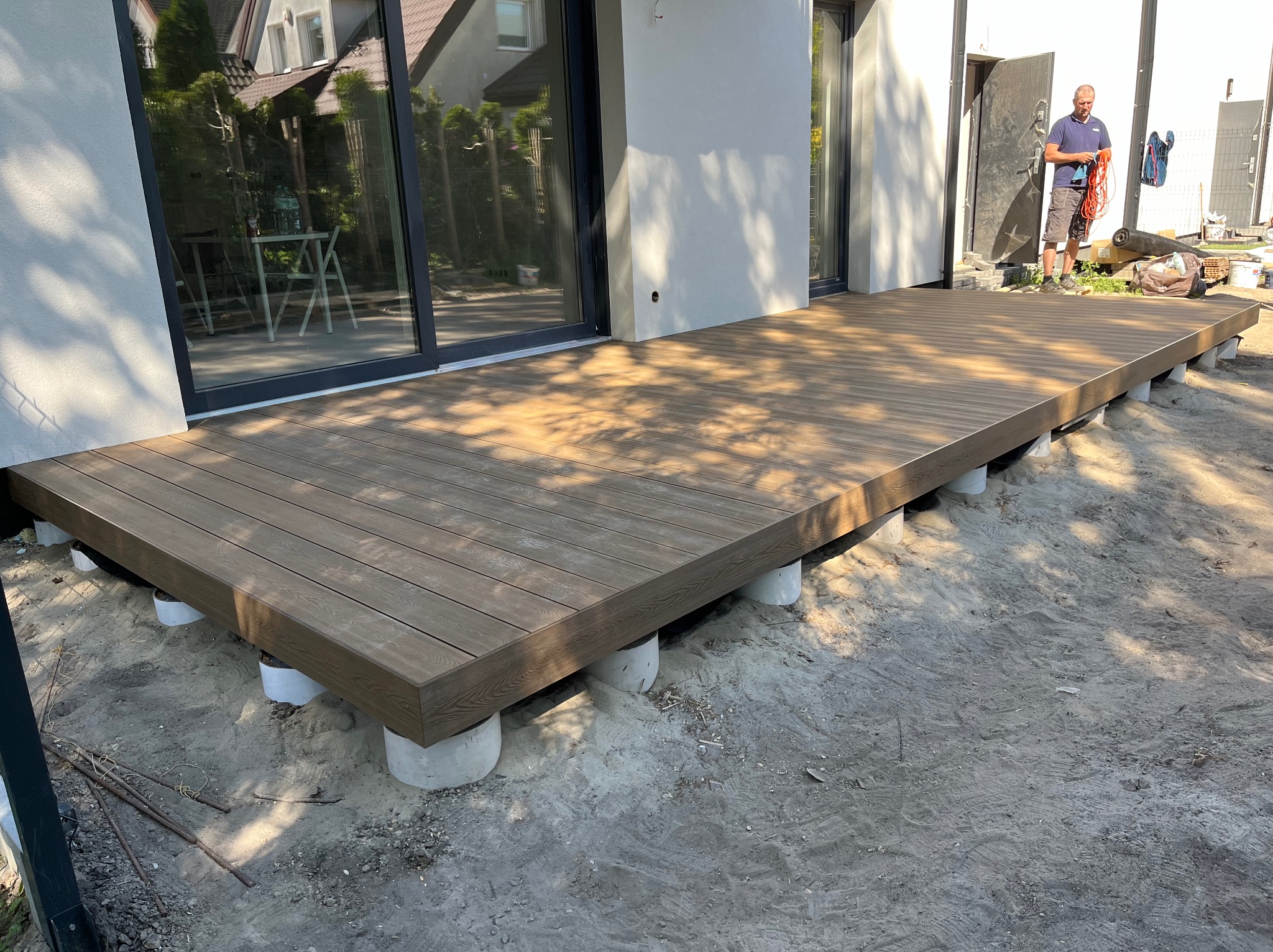 Nowoczesny taras z desek kompozytowych w odcieniach brązu, wsparty na betonowych słupkach. W tle widoczne okno tarasowe i sylwetka mężczyzny.