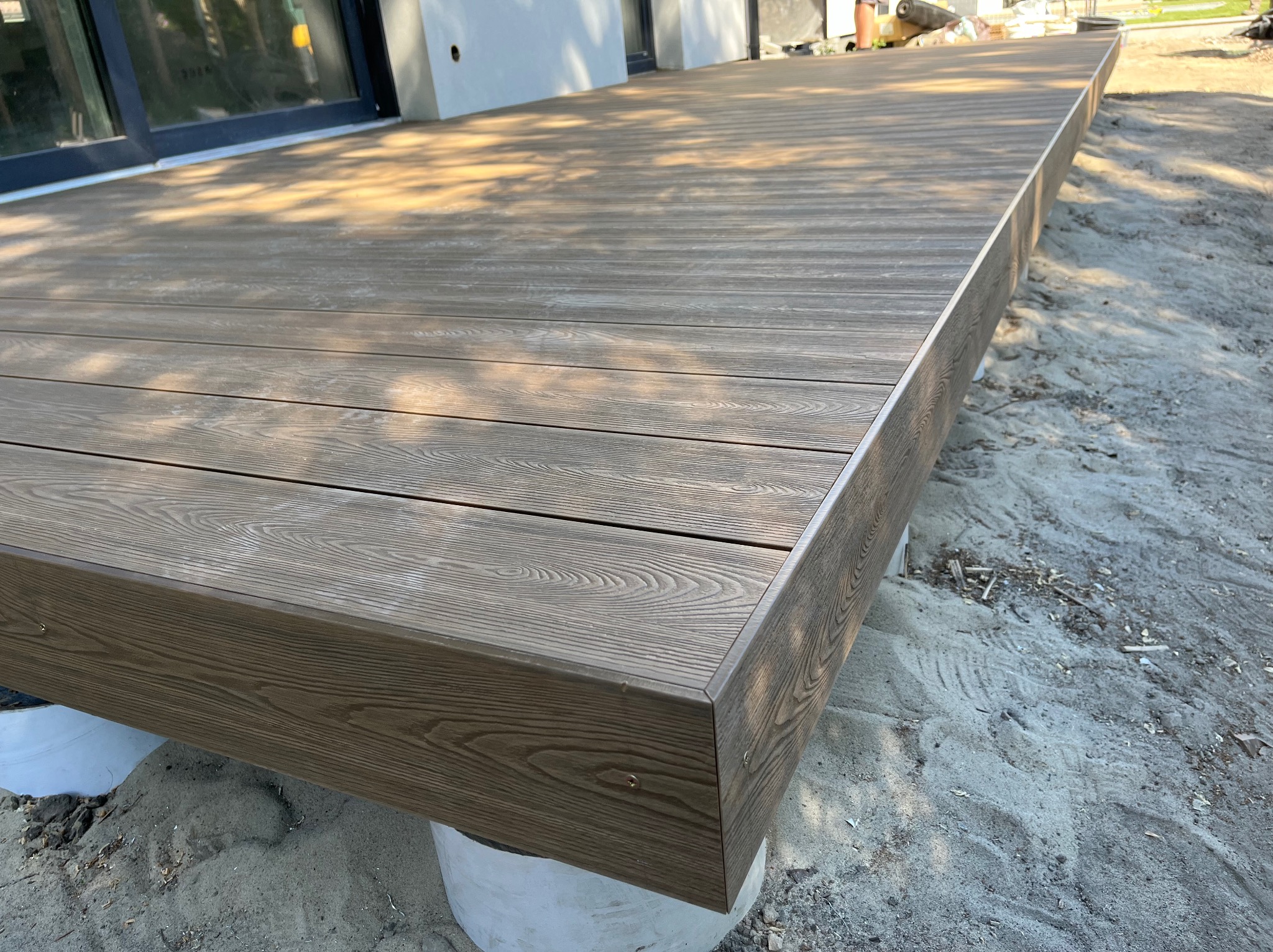 Nowoczesny taras z deski kompozytowej w odcieniach brązu, z widoczną strukturą drewna i precyzyjnym wykończeniem krawędzi. Podkonstrukcja na betonowych podporach.