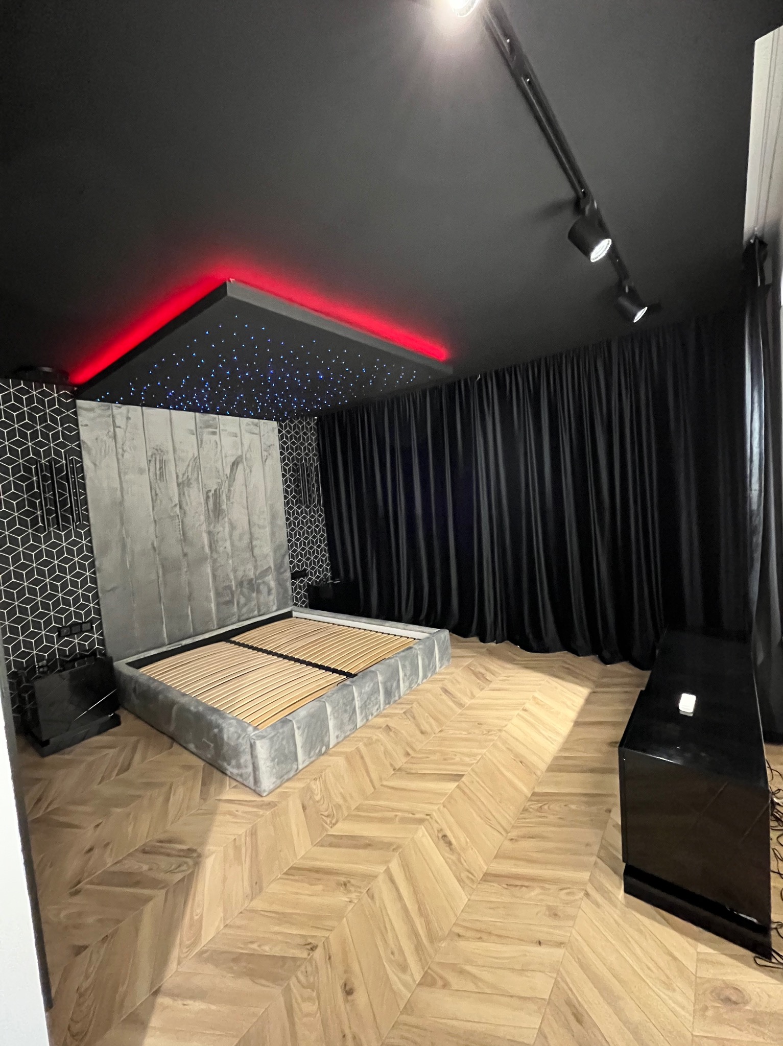 Nowoczesna sypialnia z szarym łóżkiem, parkietem w jodełkę, czarnym sufitem z oświetleniem LED i dekoracyjnymi zasłonami. Minimalistyczny design wnętrza.