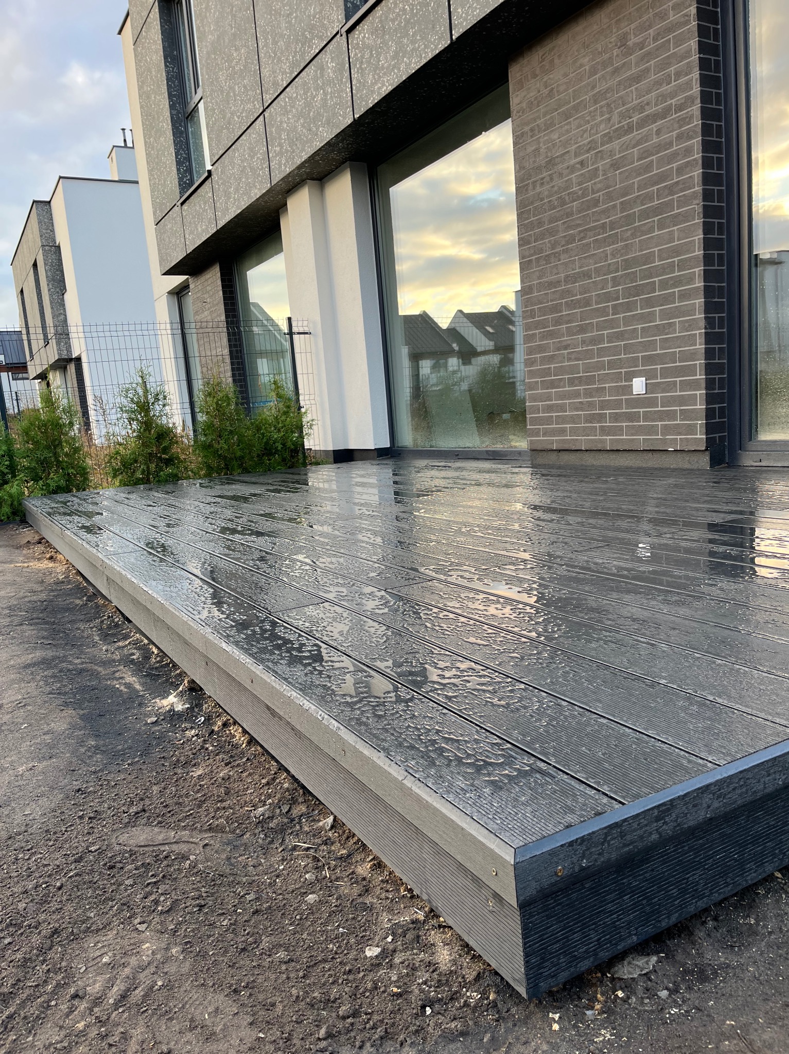 Nowoczesny taras z desek kompozytowych w kolorze grafitowym, po deszczu. Widoczne odbicia w mokrej powierzchni, minimalistyczna architektura domu w tle.