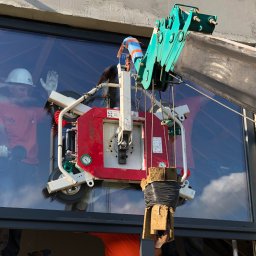 KS System KAMIL SZCZEPANIK - Montaż dużego okna z użyciem dźwigu i przyssawki, widoczny pracownik w kasku i z przyssawkami w dłoniach, odbijający się w szybie, surowy beton wokół otworu okiennego.