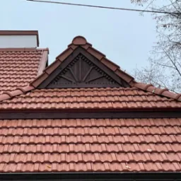 Czerwona dachówka ceramiczna na dachu domu z ozdobnym szczytem i widocznymi rynnami, na tle pochmurnego nieba i gałęzi drzew