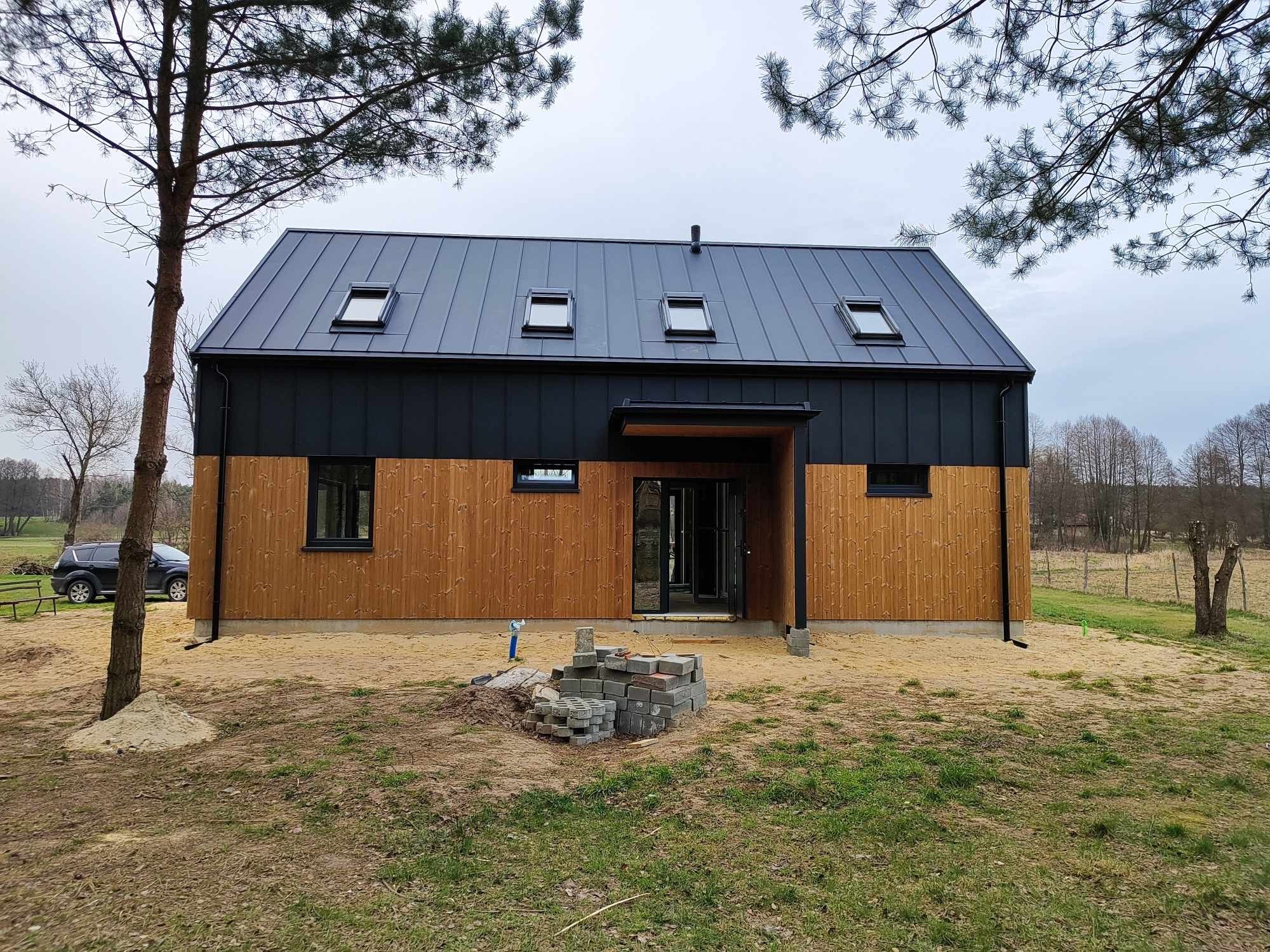 Nowoczesny dom z czarnym, metalowym dachem z czterema oknami dachowymi i drewnianą elewacją, widok z przodu, teren wokół domu w trakcie zagospodarowywania, sterta kostki brukowej przed wejściem.