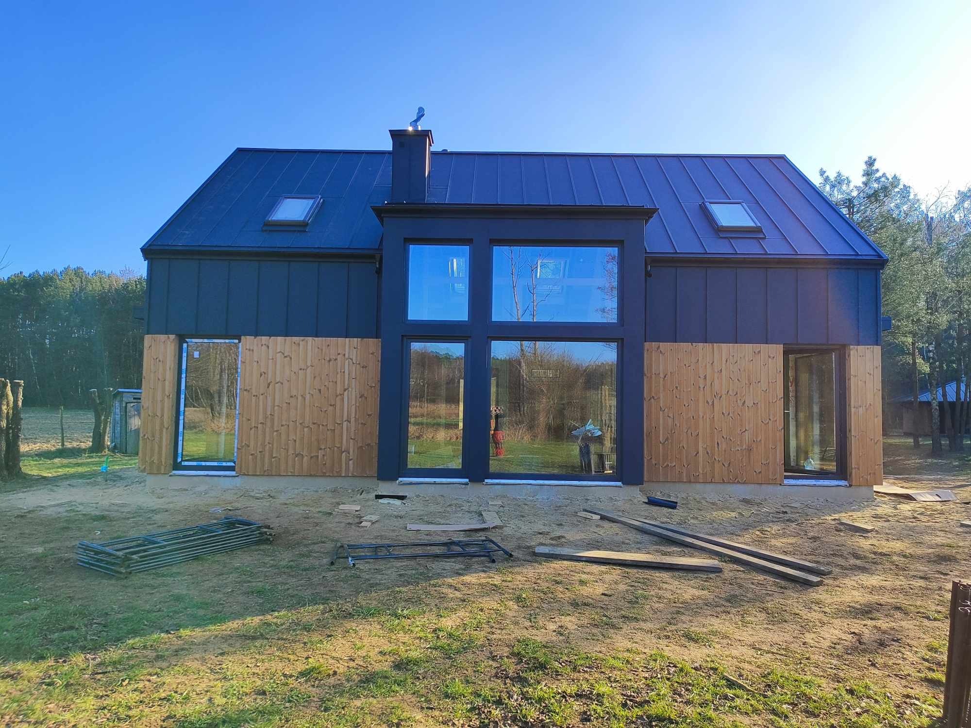 Nowoczesny dom jednorodzinny z ciemnym dachem z blachy na rąbek stojący, dużymi oknami i drewnianą elewacją w trakcie budowy, widoczne rusztowania i elementy montażowe na trawniku.