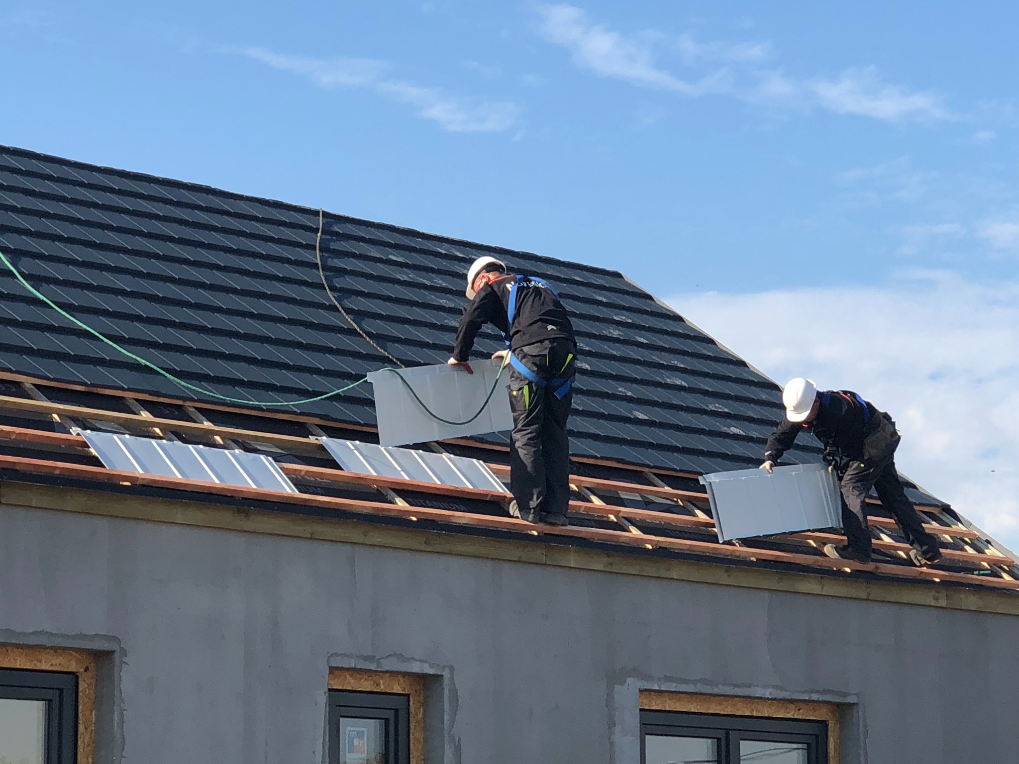 Dwóch dekarzy w uprzężach, montujących srebrne blachy na drewnianej konstrukcji dachu, z widocznymi oknami w surowej elewacji budynku.