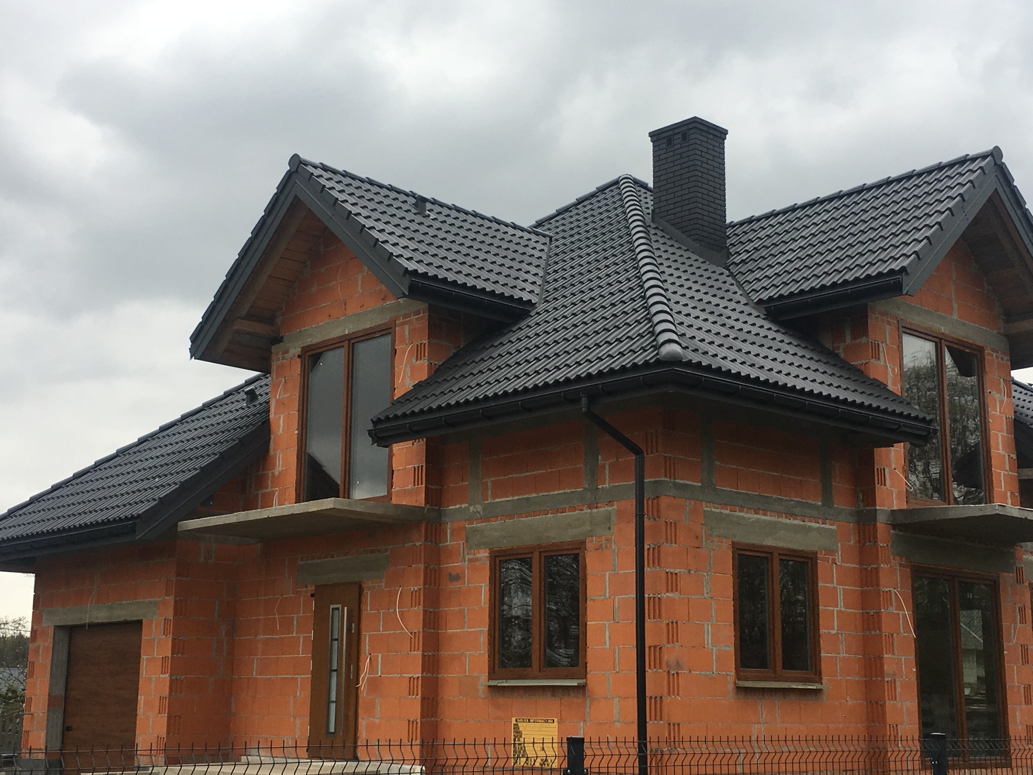 Dom w trakcie budowy z czarną dachówką, widoczne cegły i surowe wykończenie, rynny w kolorze dachu, czarny komin, okna w drewnianych ramach, pochmurne niebo.