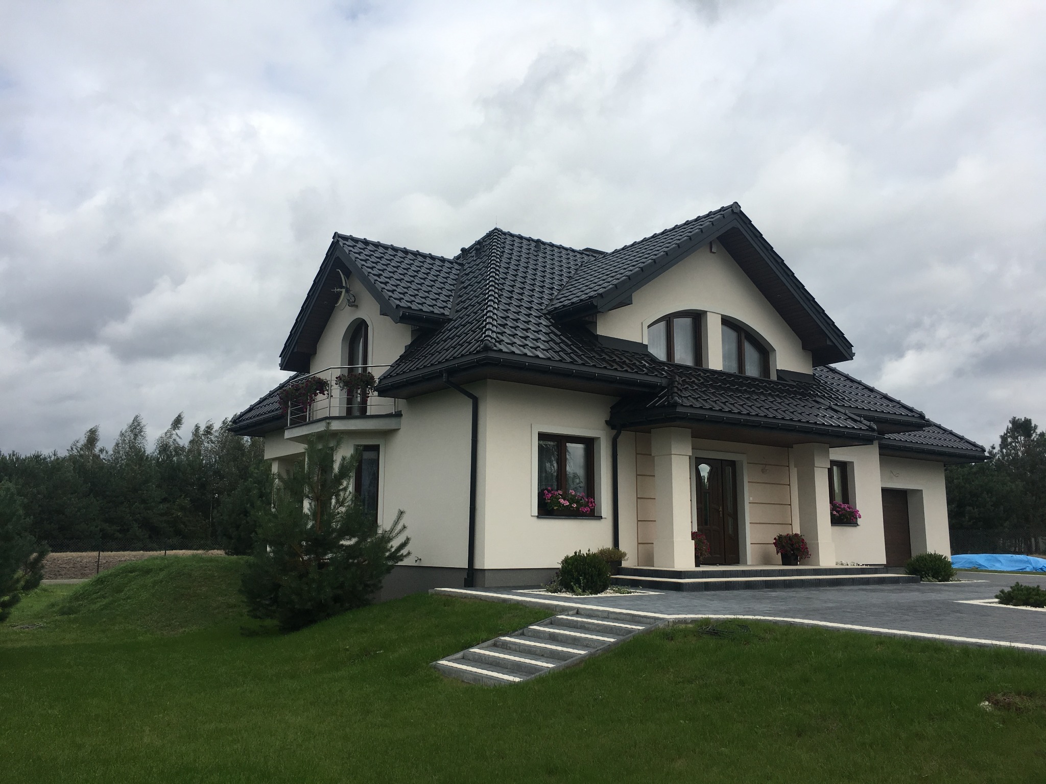 Elegancki, dwupiętrowy dom z czarnym dachem z dachówki ceramicznej, kremową elewacją, białymi kolumnami przy wejściu i zadbanym trawnikiem. Widoczne schody z kostki prowadzące do drzwi wejściowych...