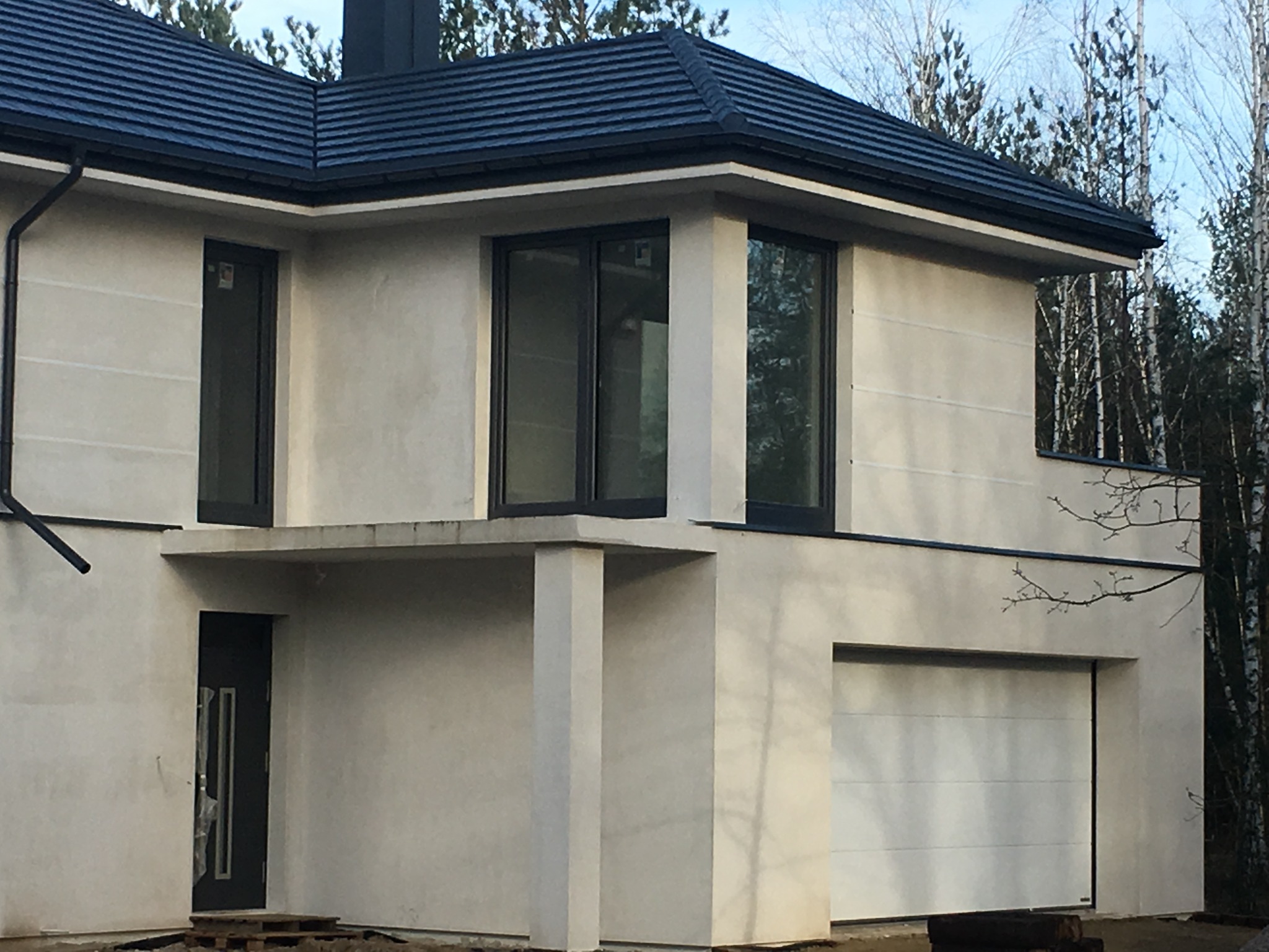 Nowoczesny dom jednorodzinny w trakcie wykańczania, z grafitową dachówką, rynnami i stolarką okienną w kolorze antracytowym, widoczna brama garażowa, okna PCV i fragment dachu z orynnowaniem.