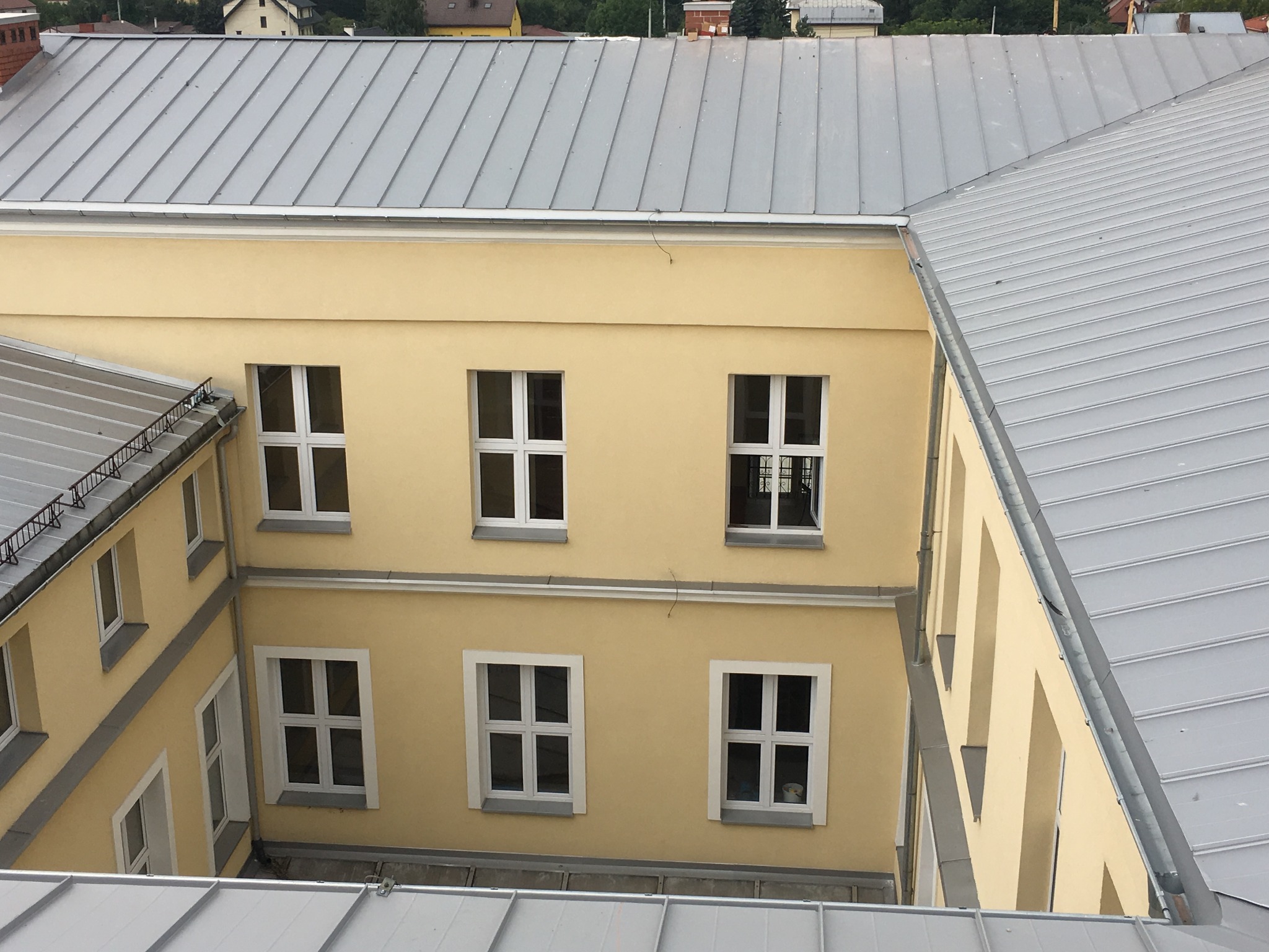 Wewnętrzny dziedziniec budynku z żółtą elewacją, widok z góry na szare, metalowe pokrycie dachowe i białe okna z podziałami.