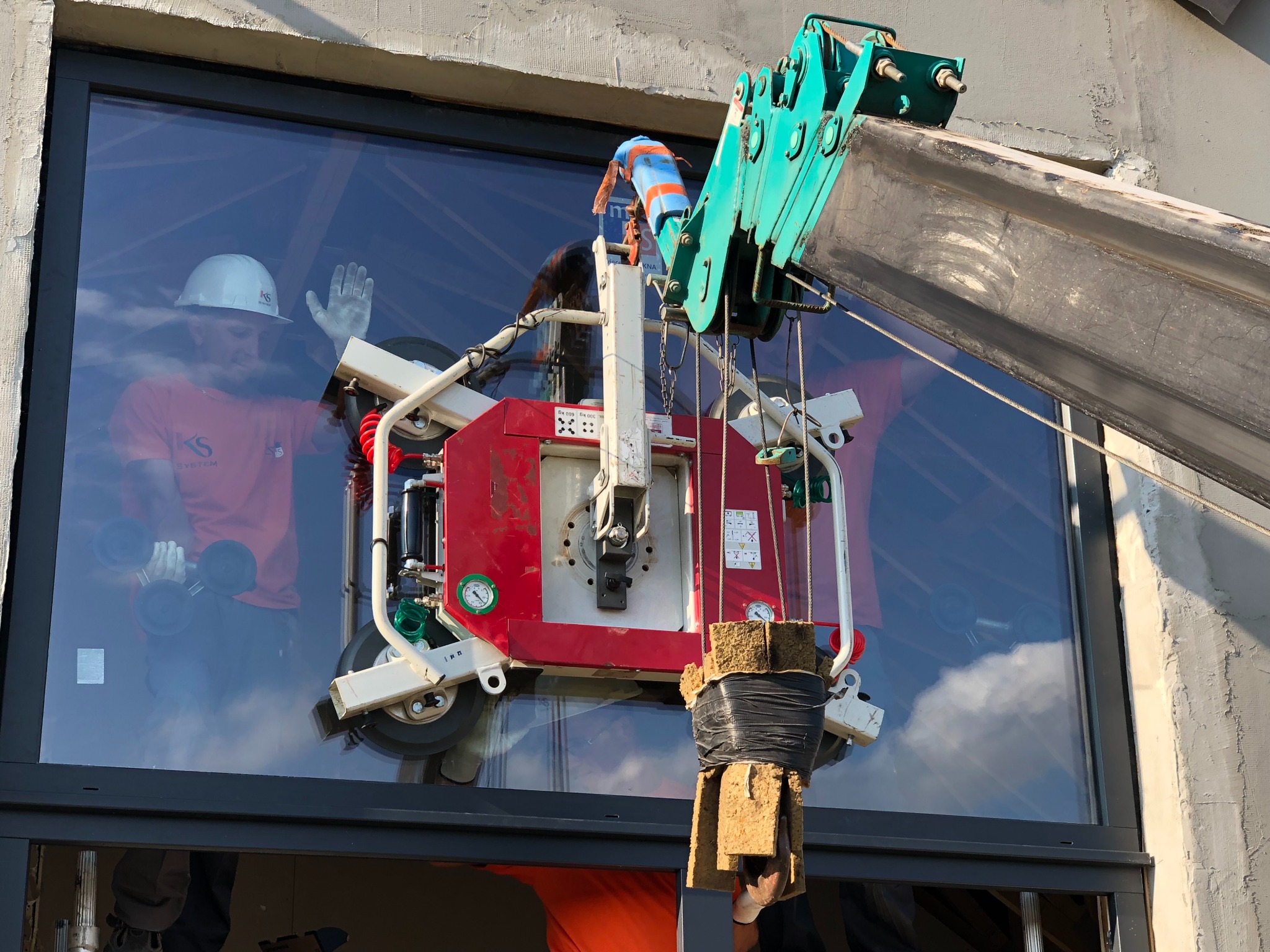 Montaż dużego okna z użyciem dźwigu i przyssawki, widoczny pracownik w kasku i z przyssawkami w dłoniach, odbijający się w szybie, surowy beton wokół otworu okiennego.