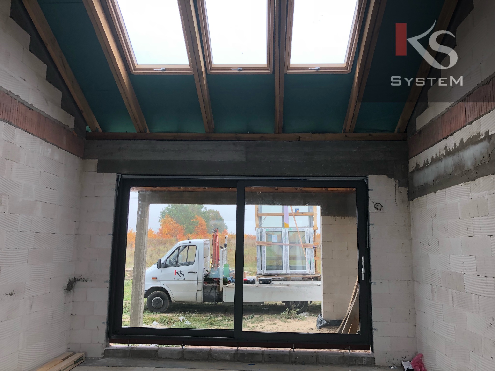 Montaż okien w surowym stanie budynku, widoczne okna połaciowe w drewnianej ramie oraz duże okno przesuwne, na zewnątrz samochód dostawczy z załadunkiem okien.