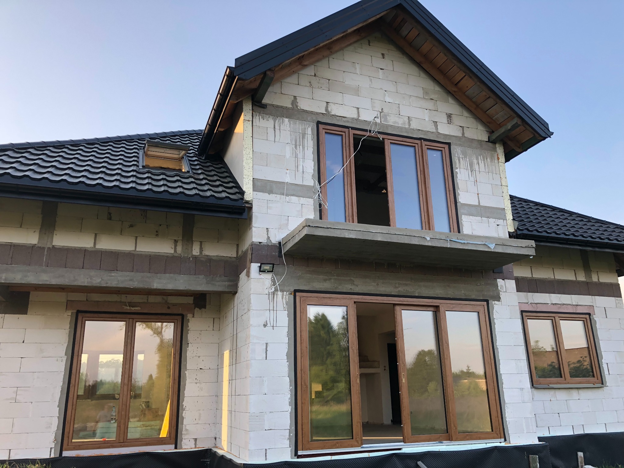 Surowy stan domu jednorodzinnego z zamontowanymi oknami z brązowymi ramami, dach pokryty ciemną dachówką, ściany z białego bloczka, widoczne elementy konstrukcyjne dachu i rynny.