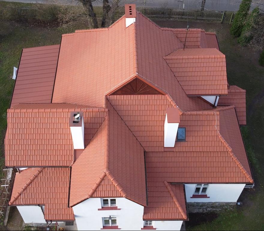 Widok z drona na dom w Garwolinie z nowym, czerwonym dachem z dachówki ceramicznej, białe kominy i okna dachowe, widoczne obróbki blacharskie na krawędziach dachu.
