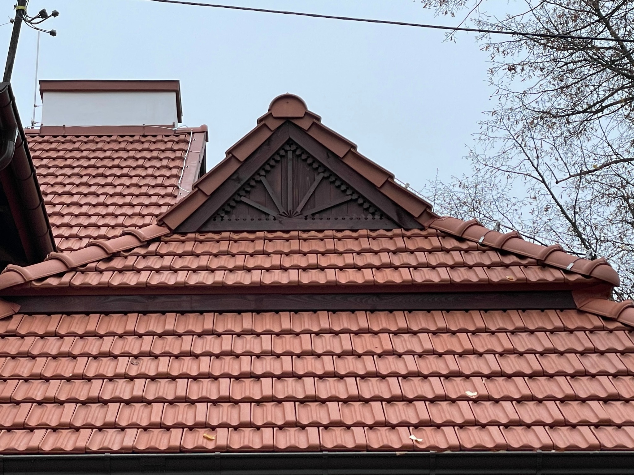 Czerwona dachówka ceramiczna na dachu domu z ozdobnym szczytem i widocznymi rynnami, na tle pochmurnego nieba i gałęzi drzew