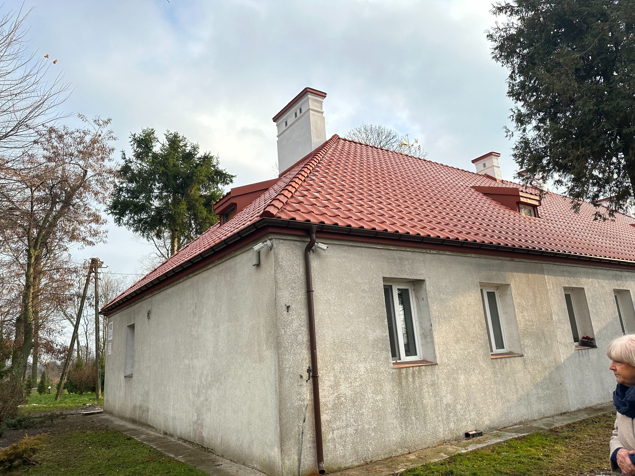 Budynek z nowym, czerwonym dachem ceramicznym, białymi kominami i rynnami, widok z zewnątrz w pochmurny dzień.