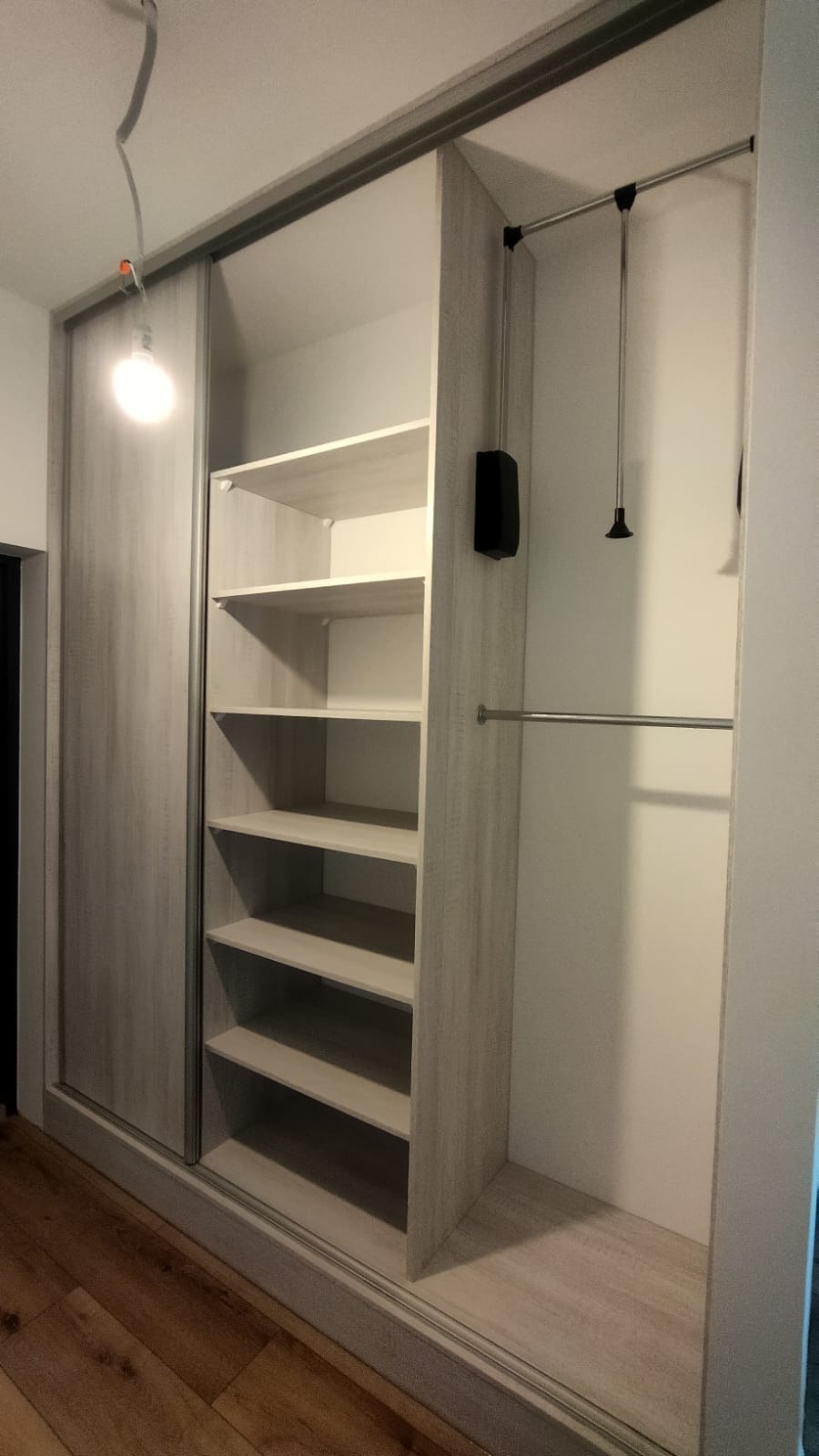 Jasna szafa wnękowa z przesuwnymi drzwiami i półkami, widoczny system drążków na ubrania. Podłoga z paneli. Ujęcie z bliska, wnętrze szafy dobrze oświetlone.
