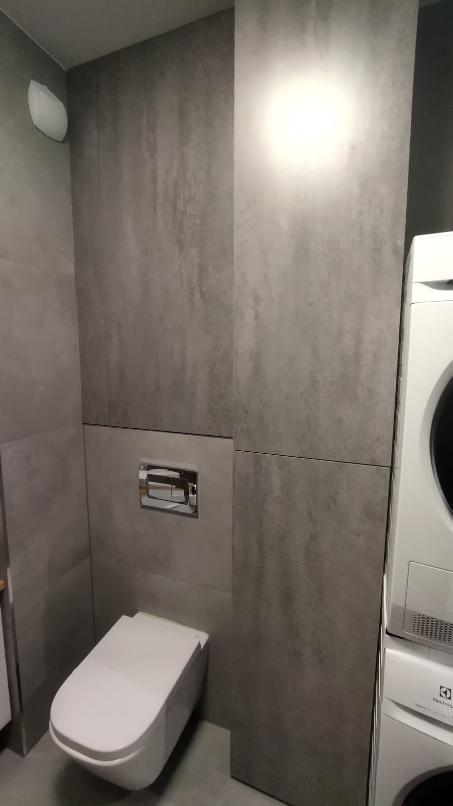 Nowoczesna toaleta z szarą, teksturowaną okładziną ścienną i podtynkową spłuczką. Obok pralka Electrolux. Minimalistyczny design, stonowana kolorystyka.