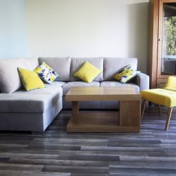 Meble Trąbka - Jasnoszara sofa narożna z żółtymi i kwiecistymi poduszkami, drewniany stolik kawowy i żółty fotel z podnóżkiem na tle drewnianej szafy z przeszklonymi drzwiami.