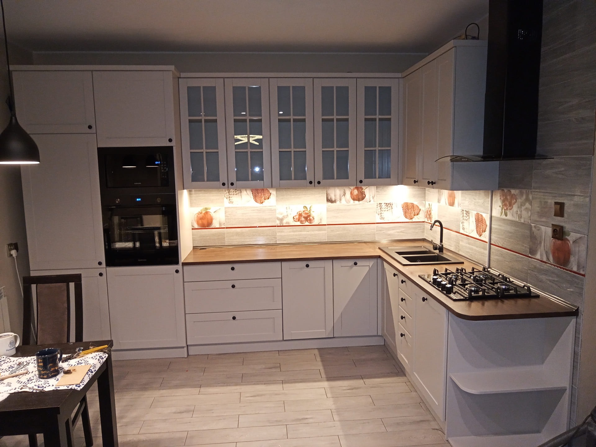 Jasna kuchnia z białymi szafkami, drewnianym blatem i płytkami z motywem owoców, oświetlona lampą wiszącą.