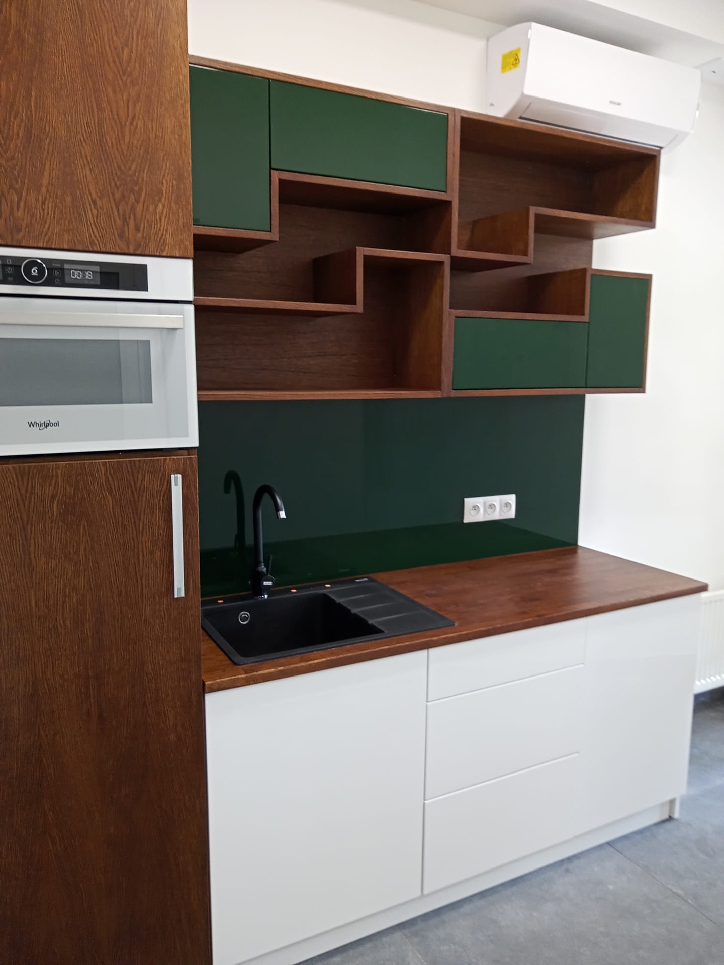 Nowoczesna kuchnia z drewnianymi szafkami górnymi i dolnymi, białymi frontami, czarnym zlewem i baterią oraz zielonymi akcentami na szafkach wiszących i ściance nad blatem.