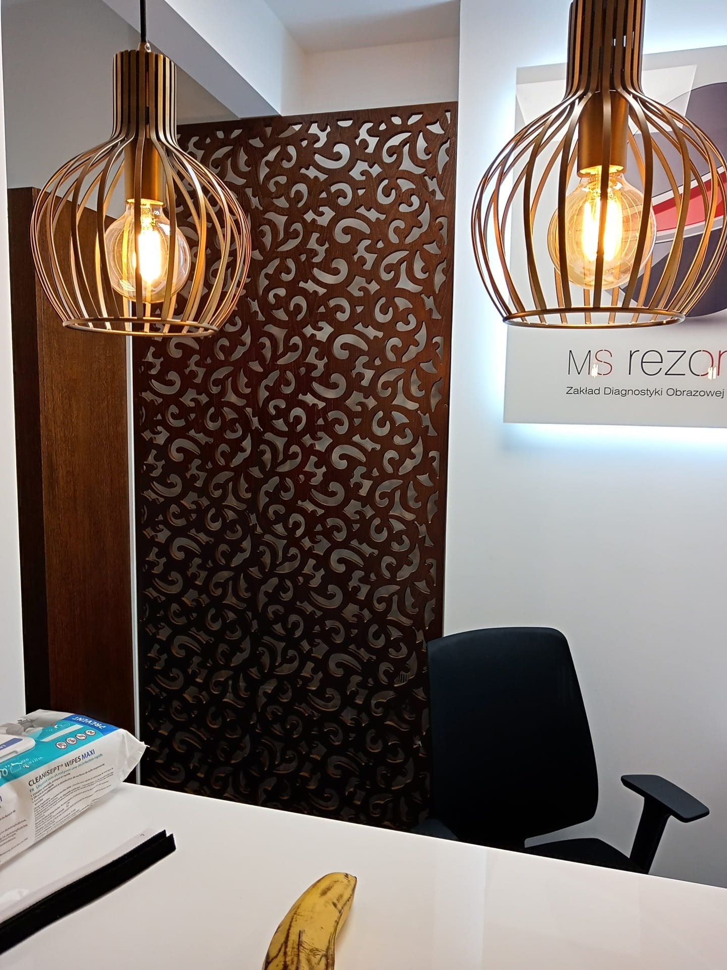 Dekoracyjna ścianka działowa z ażurowym wzorem, oświetlona wiszącymi lampami z metalowymi kloszami w kształcie klatek, widok na biurko z krzesłem i logo 'MS rezan'