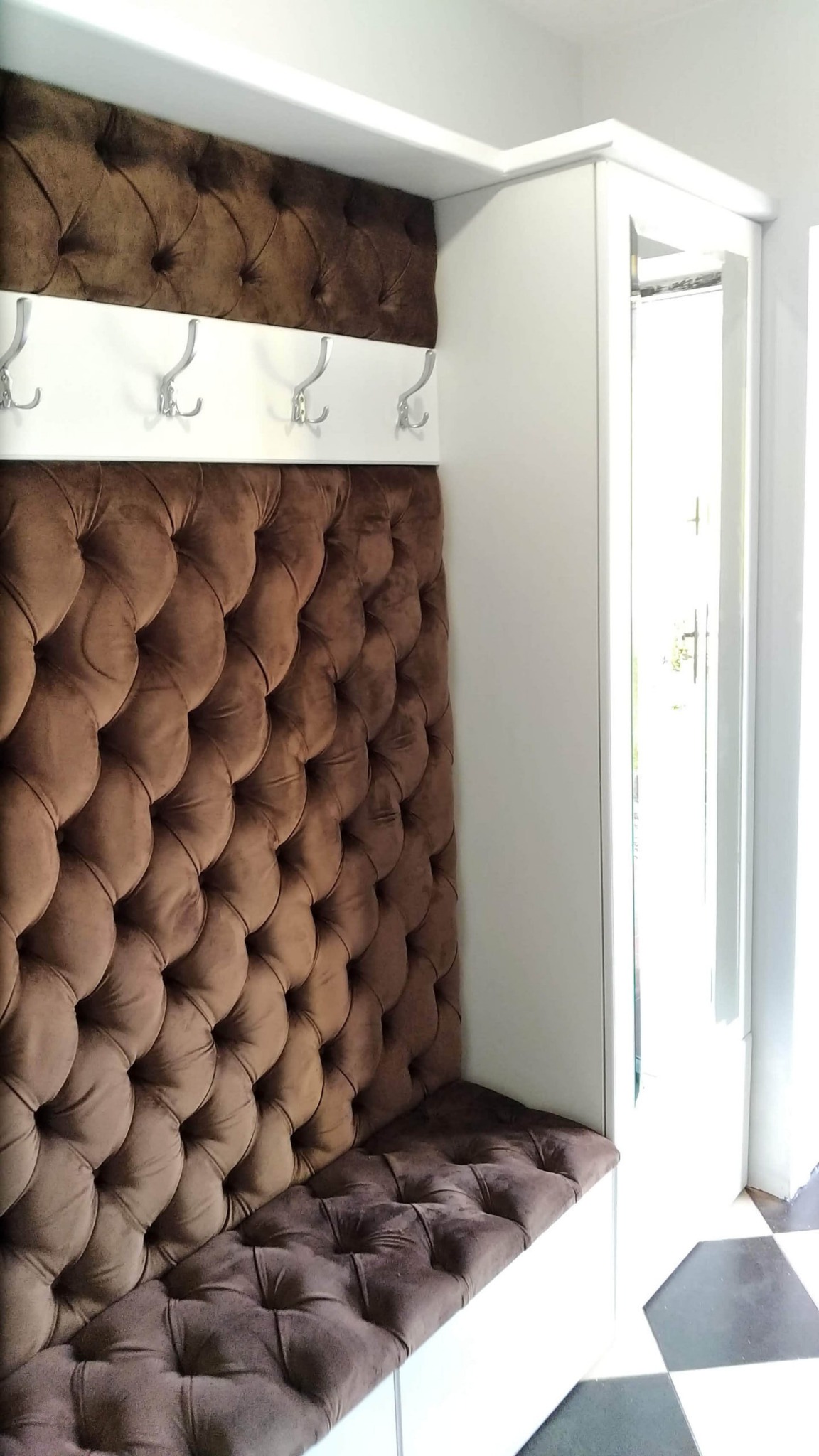 Wnęka z siedziskiem i tapicerowanym panelem ściennym w kolorze brązowym, uzupełniona o białe wieszaki i szafkę, tworząca funkcjonalną przestrzeń w przedpokoju.