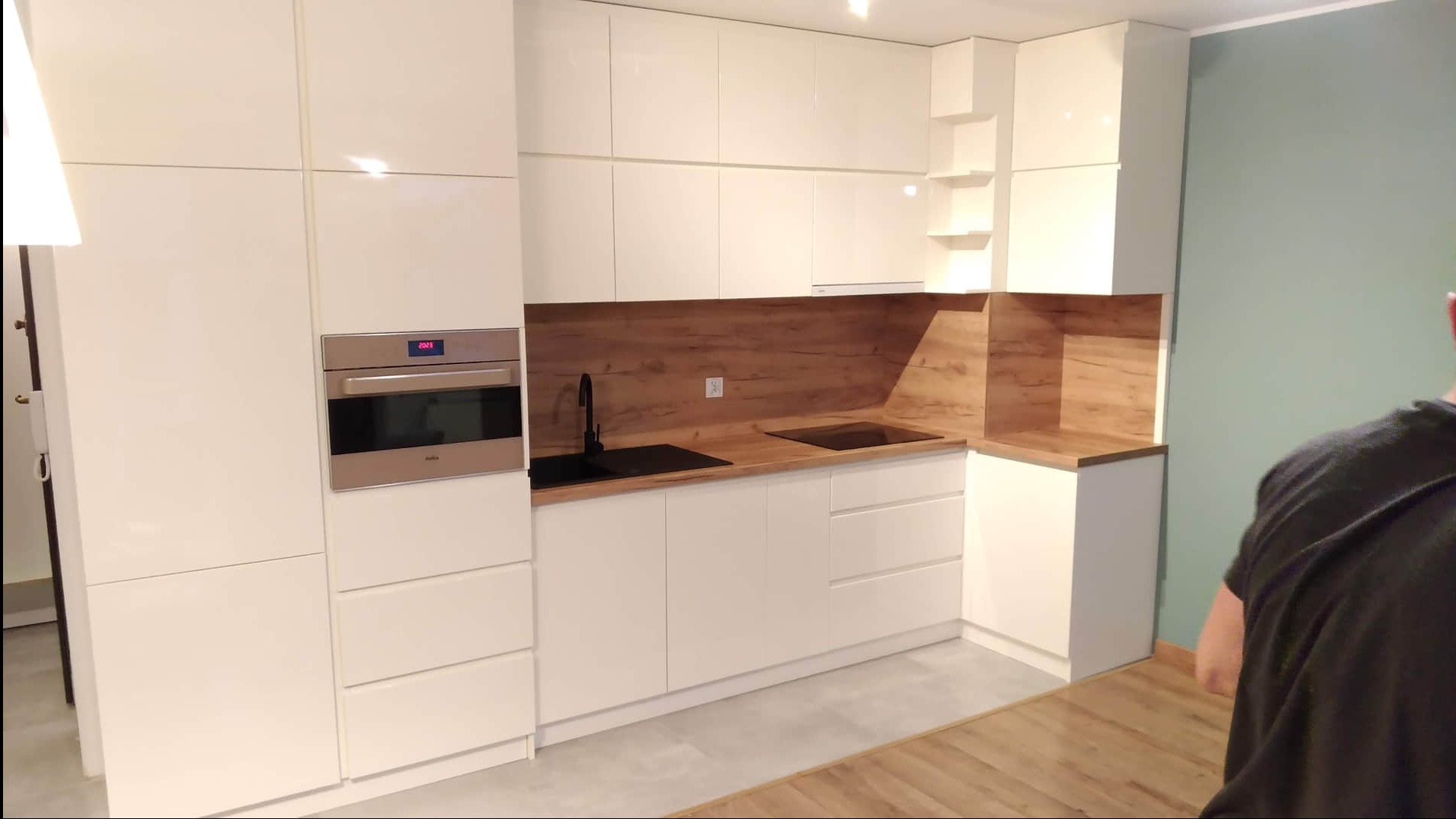 Jasna kuchnia z frontami na wysoki połysk, drewnianym blatem i panelem nad blatem. W zabudowie piekarnik z wyświetlaczem. Czarny zlew i płyta indukcyjna.