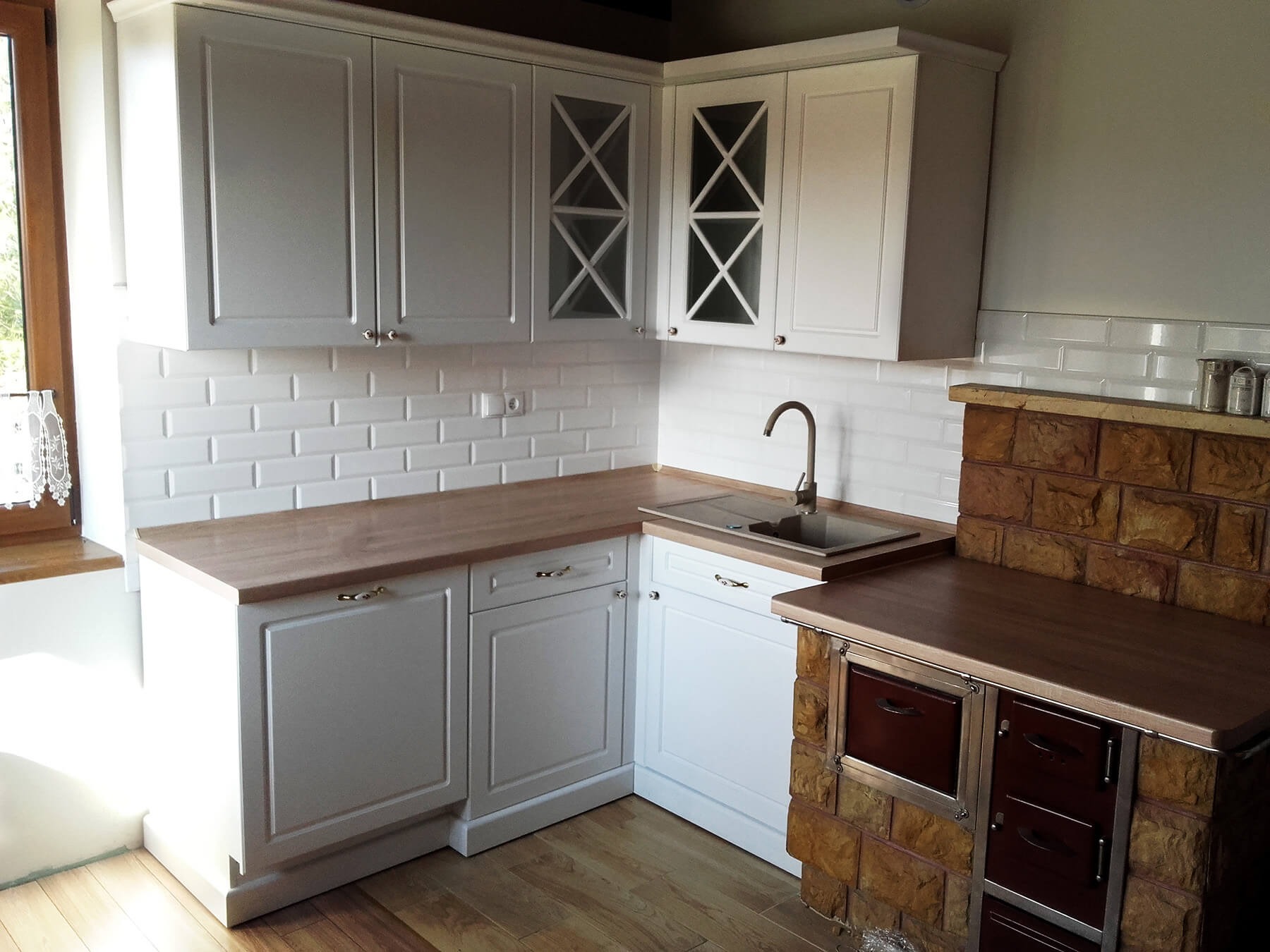 Biała kuchnia z drewnianym blatem i ceglanym piecem, płytki ścienne typu metro, szafki górne z przeszklonymi frontami w kształcie krzyża, złote uchwyty.