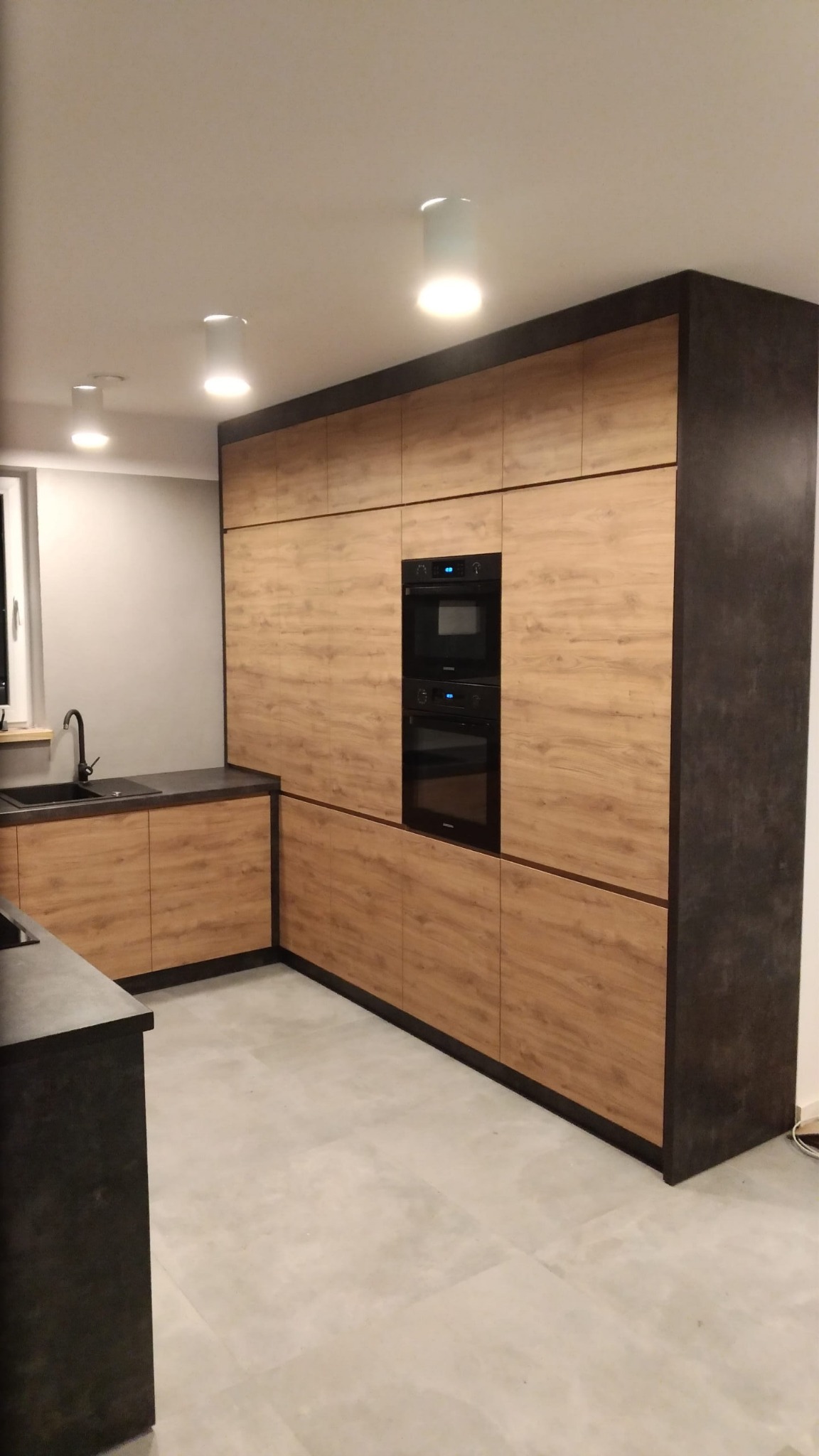 Nowoczesna zabudowa kuchenna z frontami w odcieniach drewna i ciemnoszarymi bokami, z wbudowanym piekarnikiem i mikrofalówką, oświetlona punktowymi lampami sufitowymi.
