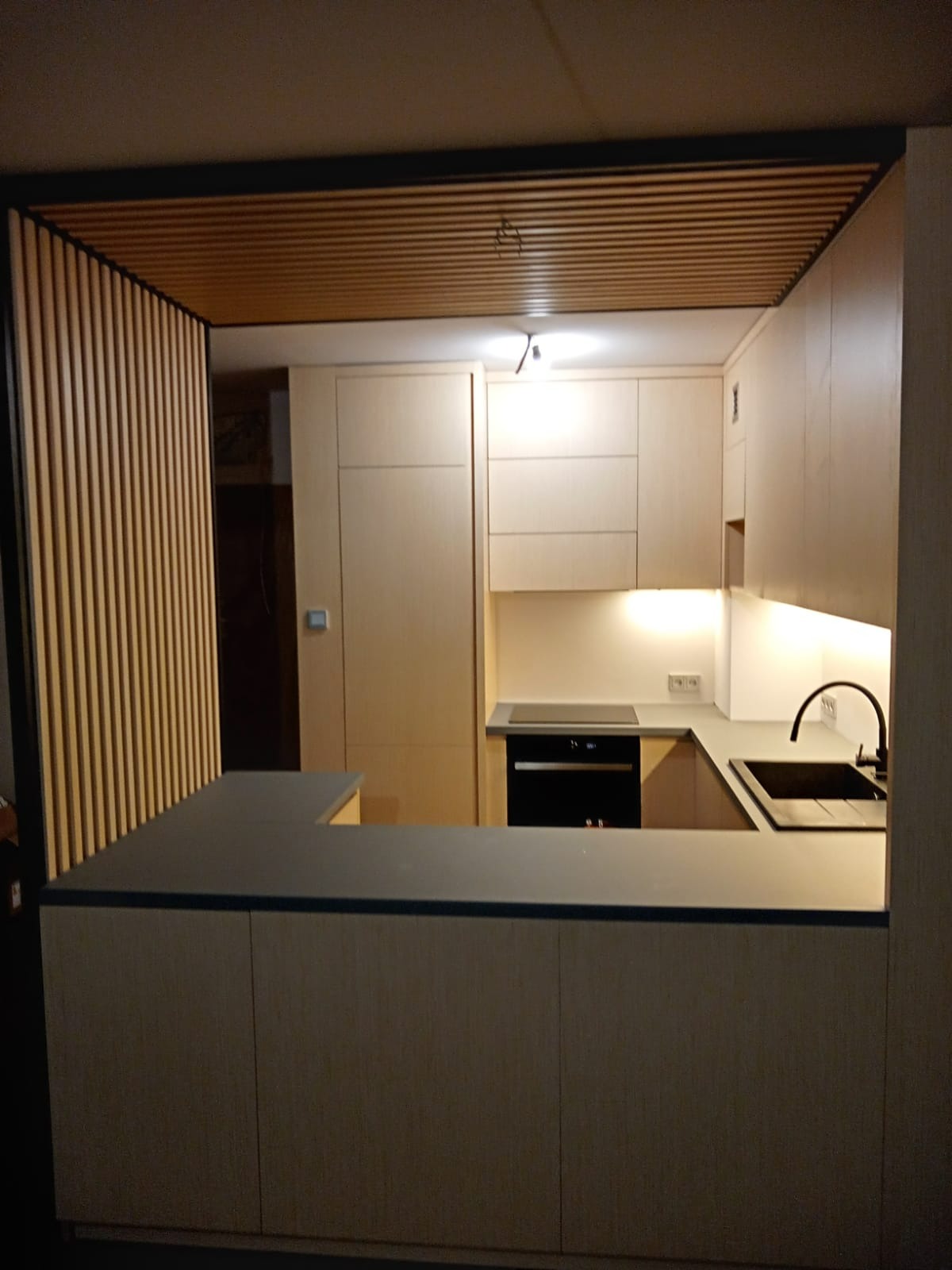 Jasna, drewniana kuchnia z półwyspem oddzielającym od reszty pomieszczenia, oświetlenie punktowe i podszafkowe, czarny zlew i bateria.