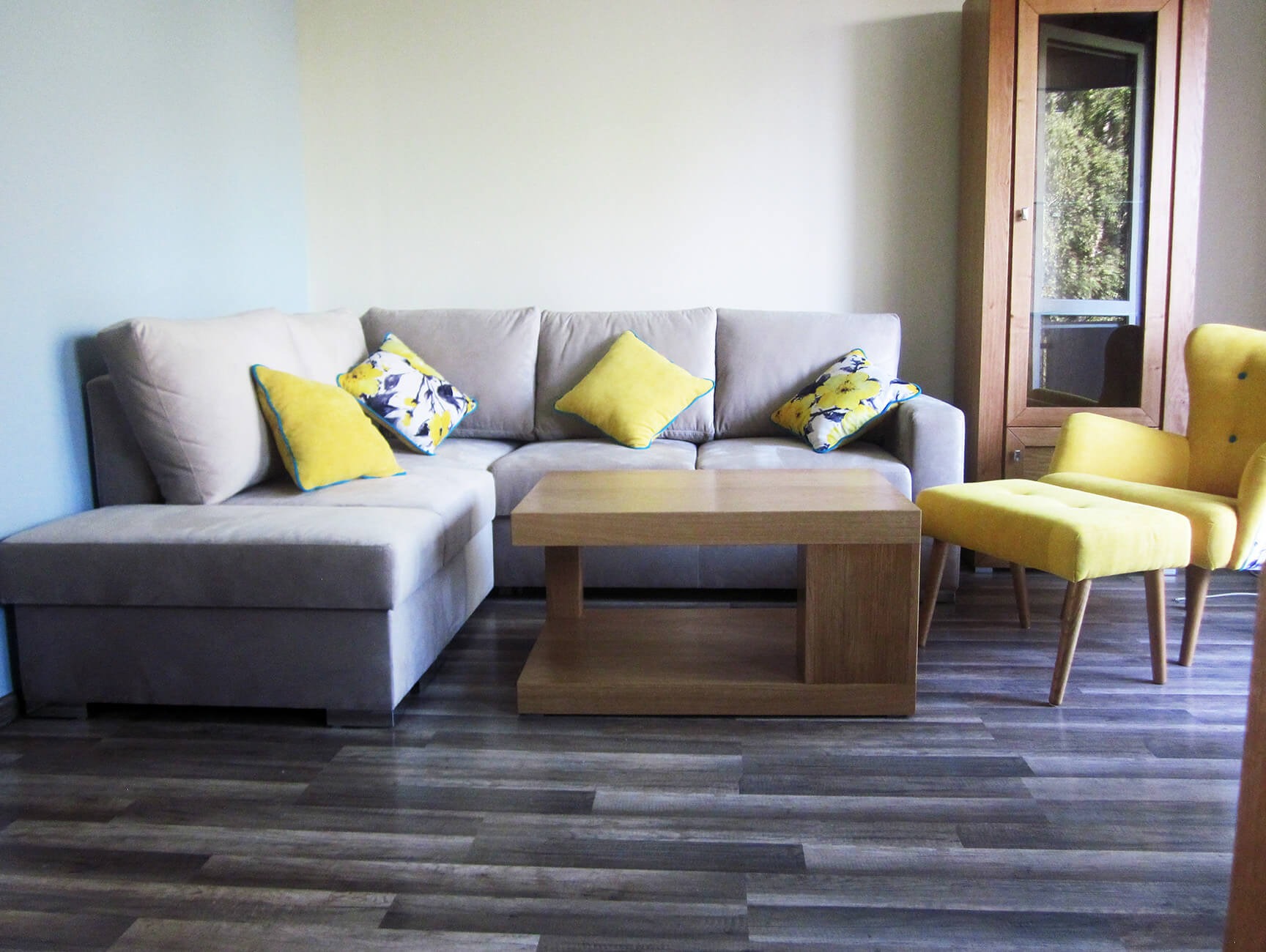 Jasnoszara sofa narożna z żółtymi i kwiecistymi poduszkami, drewniany stolik kawowy i żółty fotel z podnóżkiem na tle drewnianej szafy z przeszklonymi drzwiami.