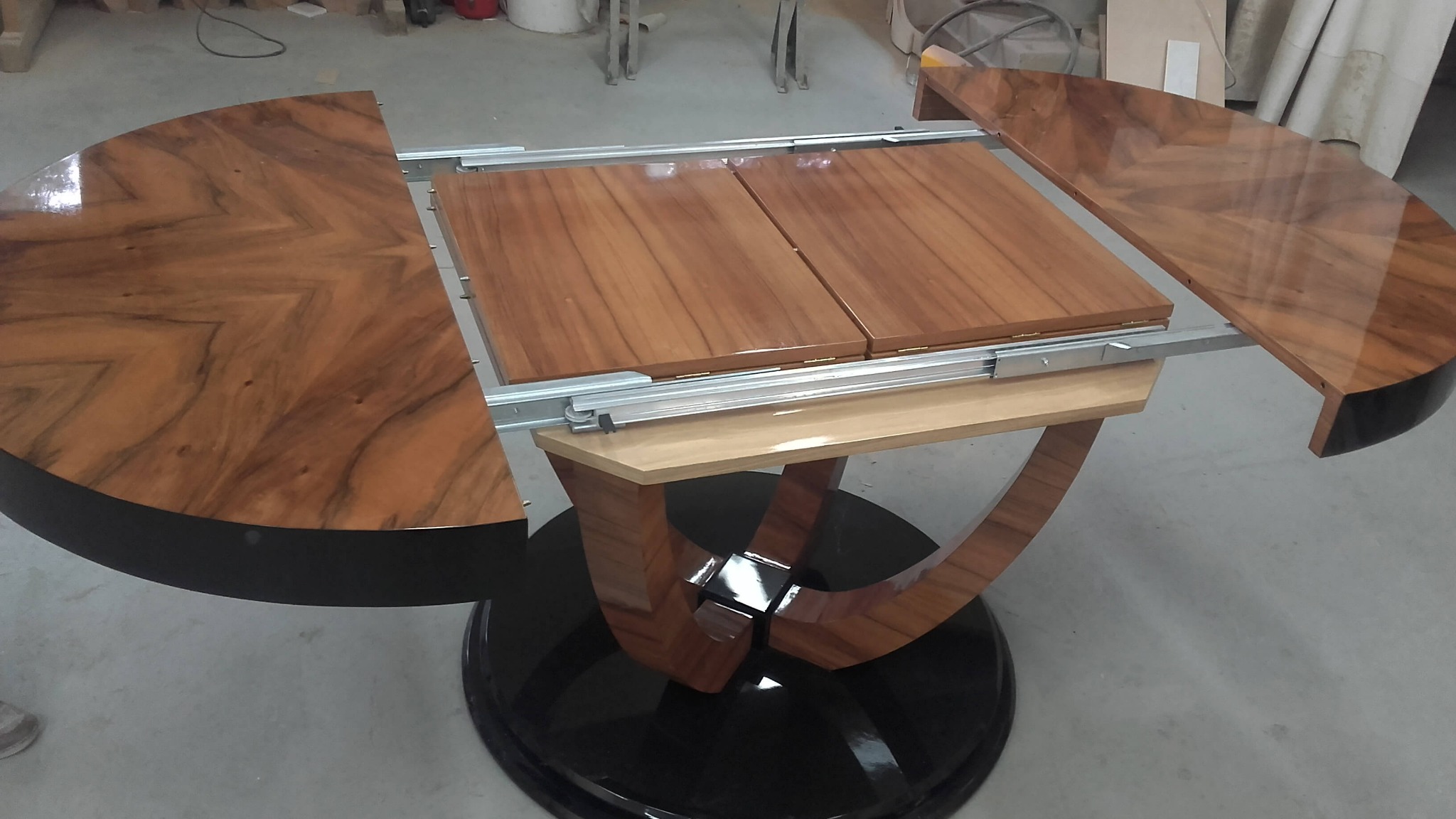 Rozkładany okrągły stół z fornirem o wzorze chevron, na czarnej, okrągłej podstawie, mechanizm rozkładania widoczny.