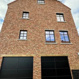 Fasada budynku z czerwonej cegły z czarnymi bramami garażowymi i oknami z ramami w tym samym kolorze, odbijającymi błękit nieba z chmurami.