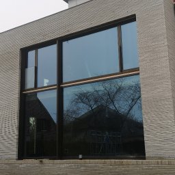 MJSCALE - Duże okno z ciemną ramą w surowym budynku z cegły. W szybach odbija się drzewo i budynek. U dołu ziemia i palety, co sugeruje etap budowy.