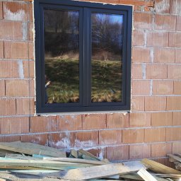 MJSCALE - Ciemne, dwuskrzydłowe okno w surowej, ceglanej ścianie budynku w trakcie budowy. Widoczne deski i zaprawa murarska. W szybie odbija się otoczenie.