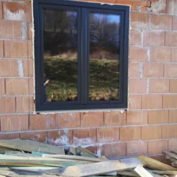 Ciemne, dwuskrzydłowe okno w surowej, ceglanej ścianie budynku w trakcie budowy. Widoczne deski i zaprawa murarska. W szybie odbija się otoczenie.