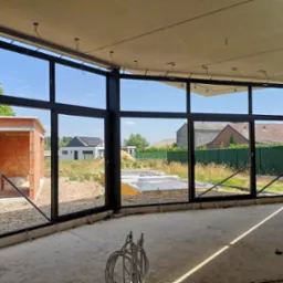 Wnętrze w stanie surowym z zamontowanymi, wielkoformatowymi oknami z czarnymi ramami. Widok na zewnątrz, gdzie widać budynki i zieleń. Surowy betonowy strop.