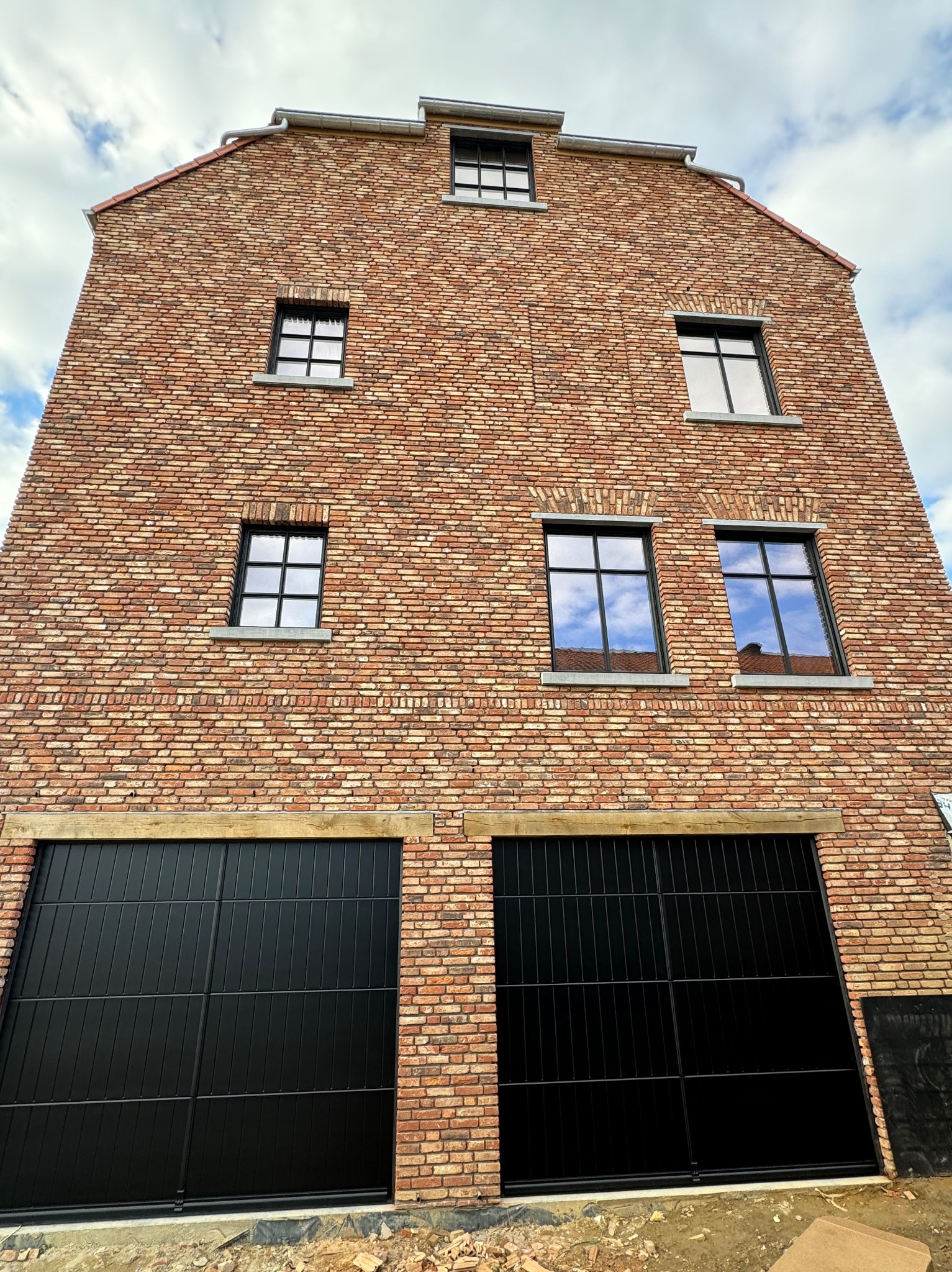 Fasada budynku z czerwonej cegły z czarnymi bramami garażowymi i oknami z ramami w tym samym kolorze, odbijającymi błękit nieba z chmurami.