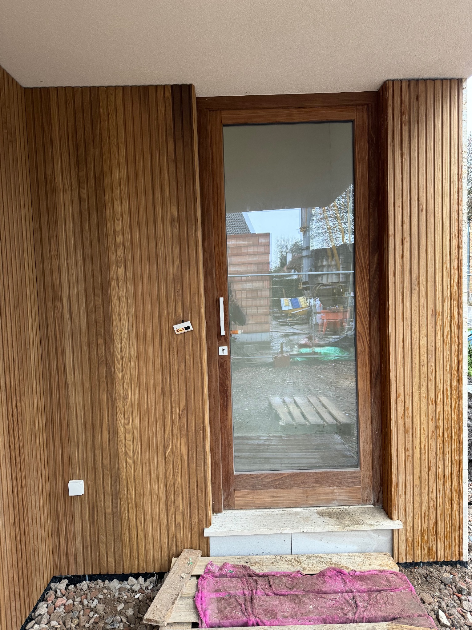 Drewniane drzwi zewnętrzne z szybą, wkomponowane w elewację z paneli drewnianych. Widoczne detale wykończenia, surowy teren budowy i odbicie otoczenia w szybie.
