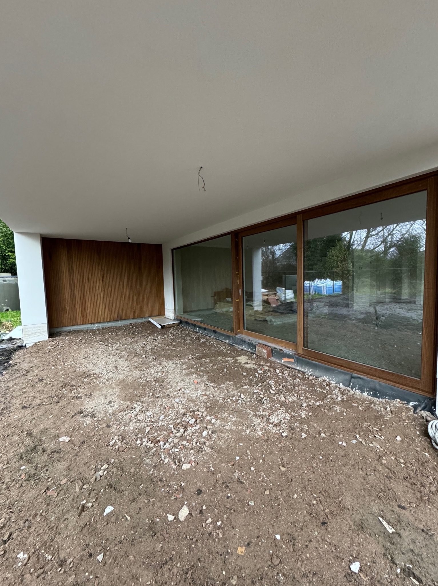 Montaż dużych, przesuwnych okien z drewnianymi ramami w nowoczesnym domu. Widoczny surowy teren przed oknami i drewniana boazeria na ścianie.