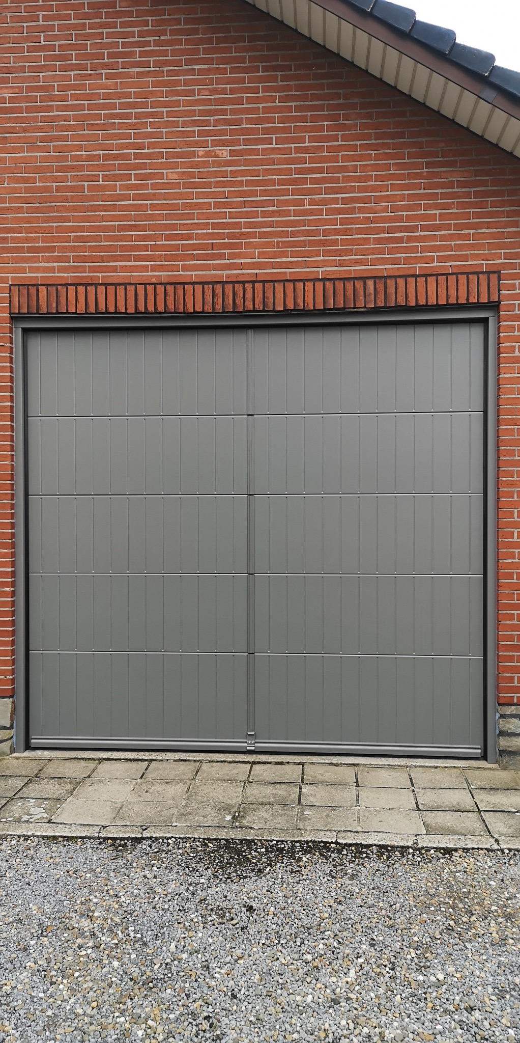 Szara segmentowa brama garażowa z pionowymi panelami, zamontowana w czerwonej ceglanej ścianie. Widok na zewnątrz, kostka brukowa i żwir przed bramą.