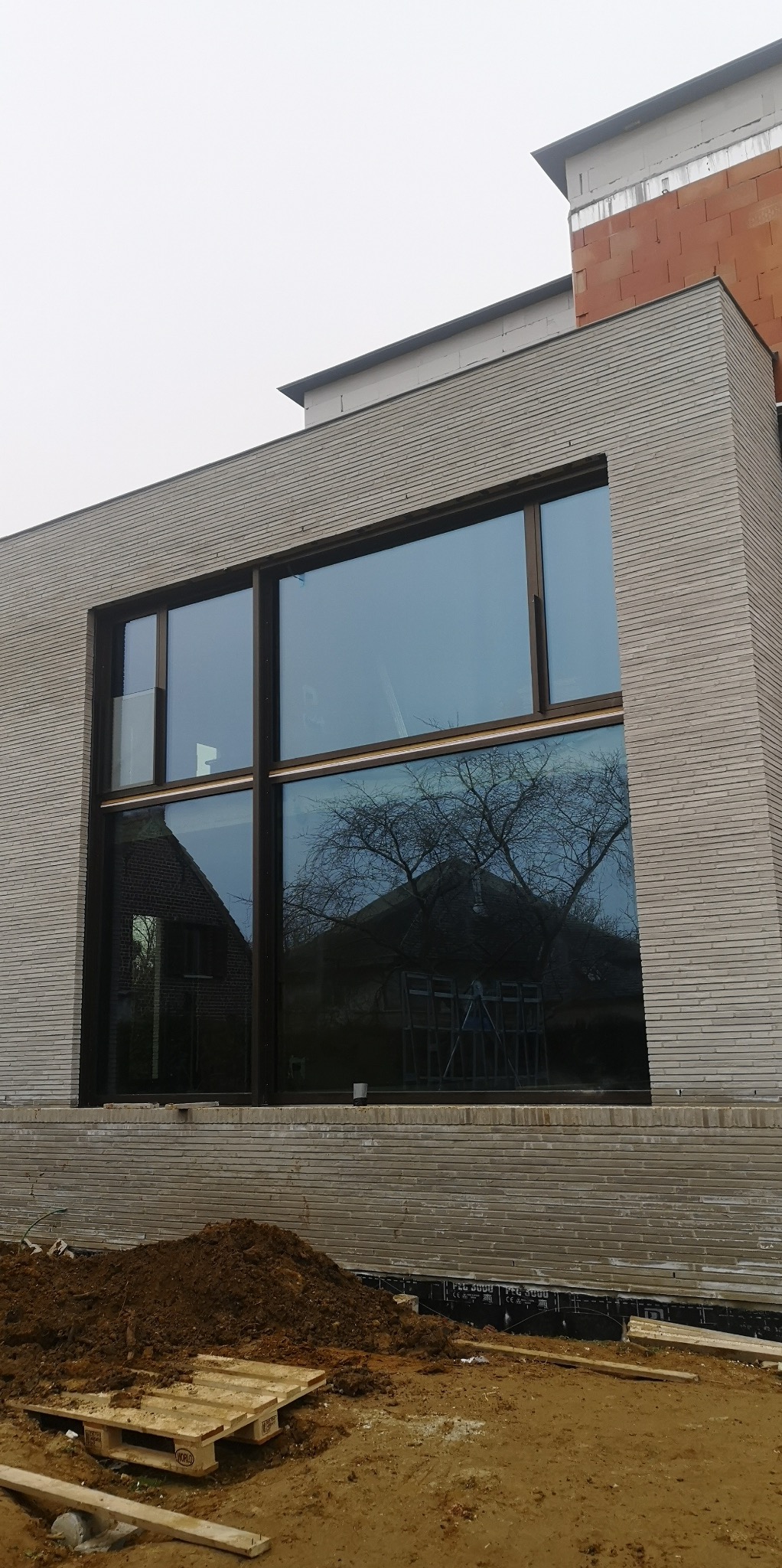 Duże okno z ciemną ramą w surowym budynku z cegły. W szybach odbija się drzewo i budynek. U dołu ziemia i palety, co sugeruje etap budowy.