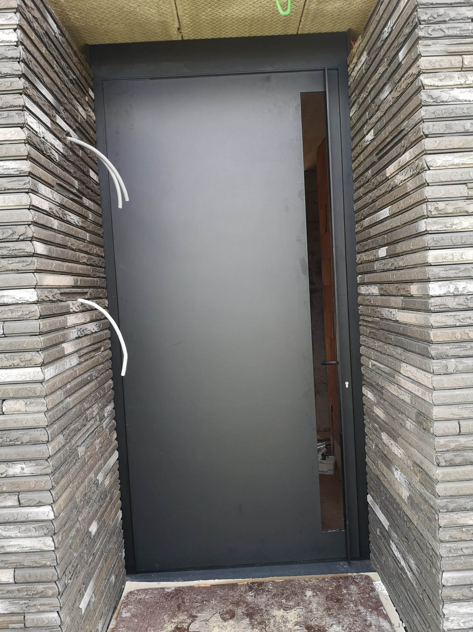 Nowoczesne, grafitowe drzwi zewnętrzne w surowej, ceglanej elewacji. Widoczne przewody elektryczne i fragment wnętrza przez przeszklenie.