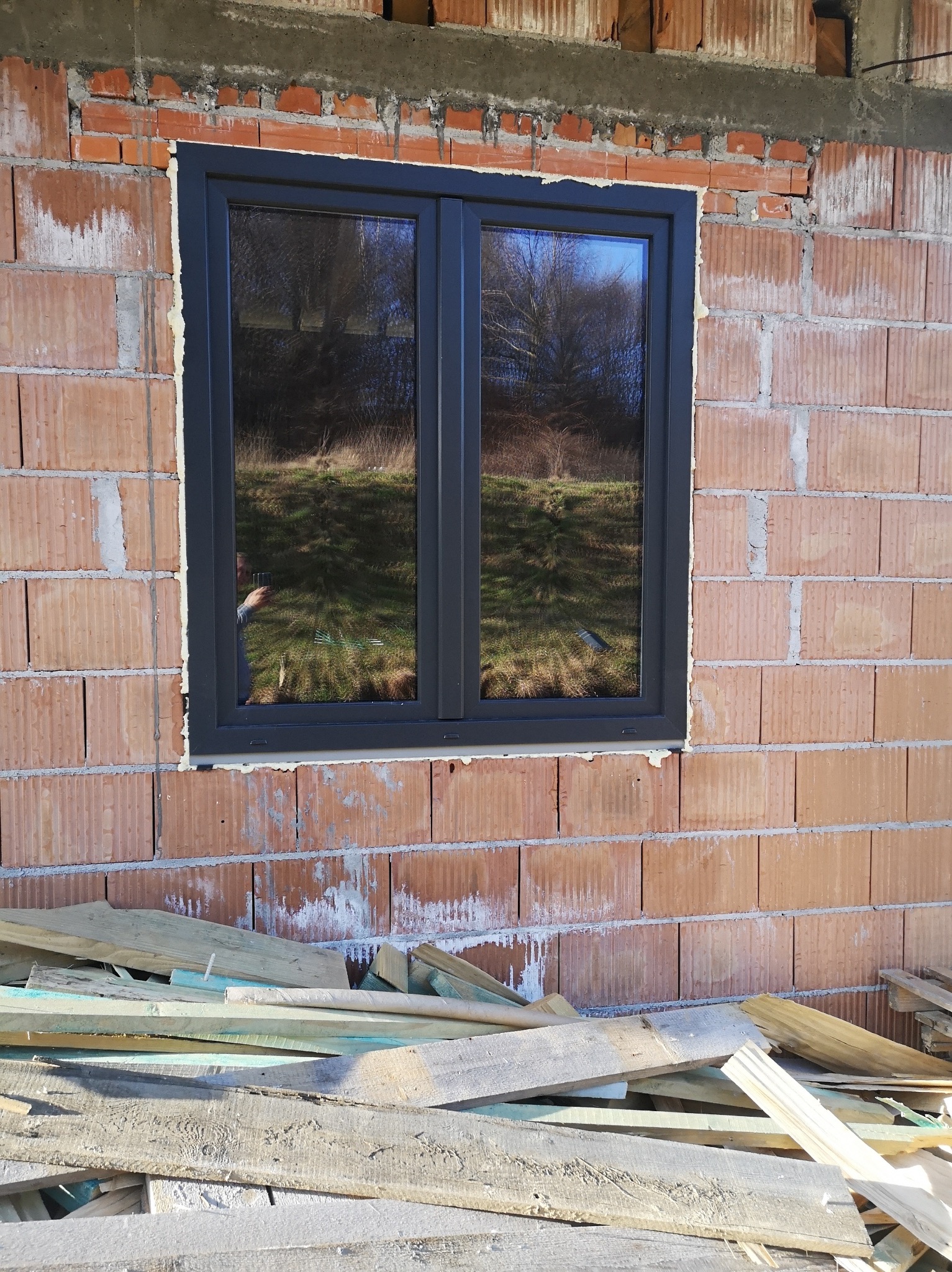 Ciemne, dwuskrzydłowe okno w surowej, ceglanej ścianie budynku w trakcie budowy. Widoczne deski i zaprawa murarska. W szybie odbija się otoczenie.