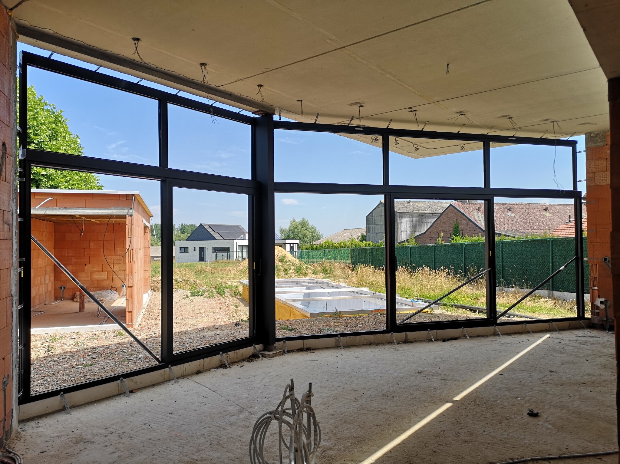 Wnętrze w stanie surowym z zamontowanymi, wielkoformatowymi oknami z czarnymi ramami. Widok na zewnątrz, gdzie widać budynki i zieleń. Surowy betonowy strop.