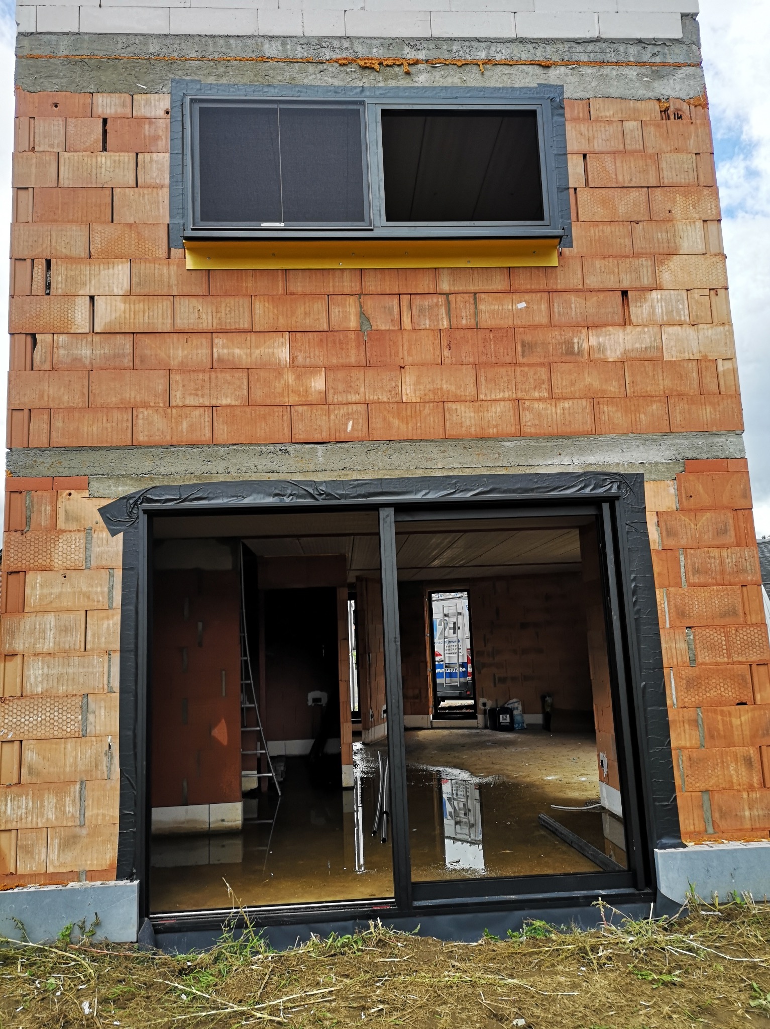 Surowy mur z zamontowanym oknem i drzwiami przesuwnymi z czarnymi ramami. Wewnątrz woda i drabina. Widok z zewnątrz na budowę w trakcie prac.