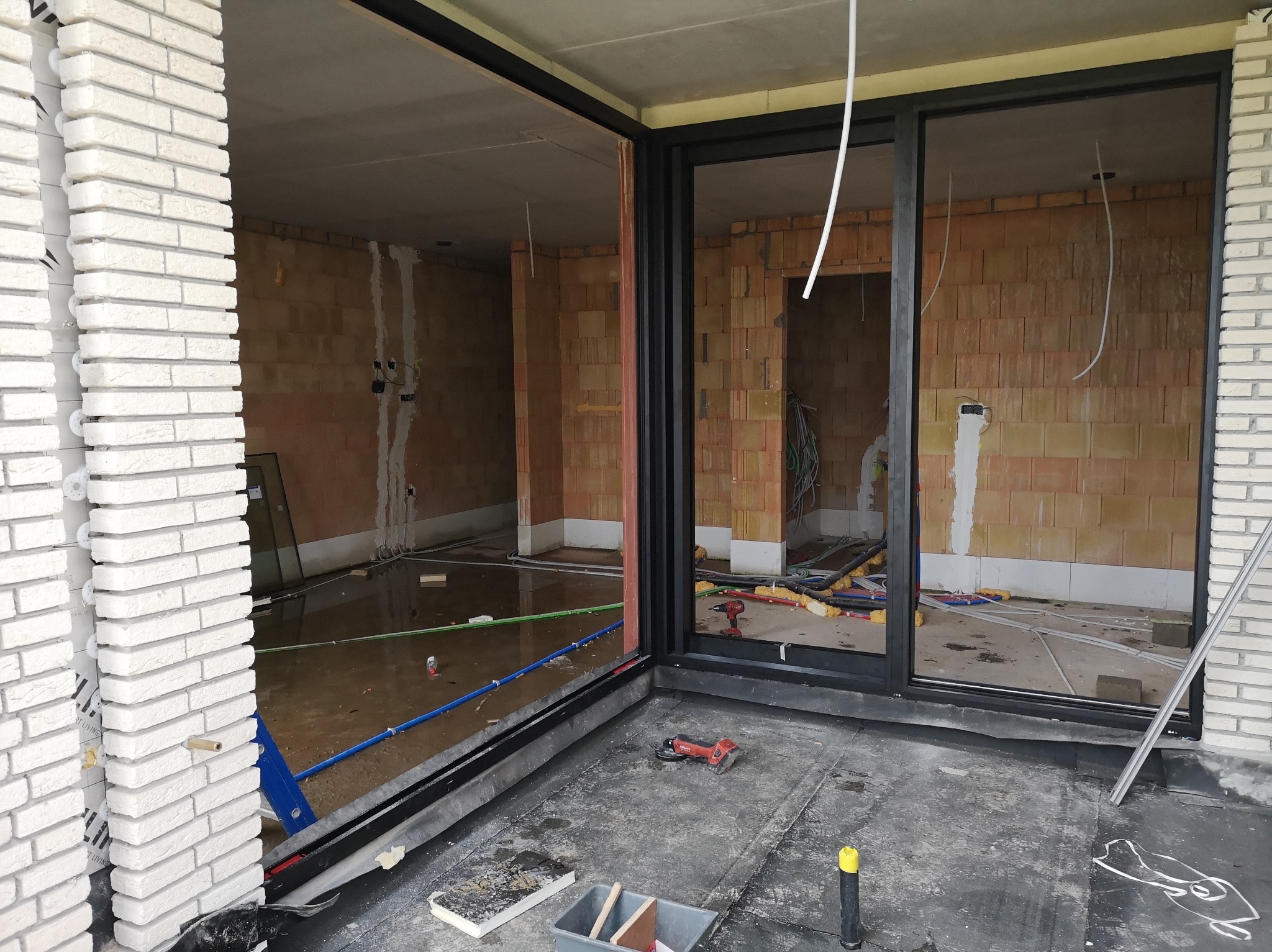 Montaż dużych, przesuwnych drzwi z czarnymi ramami w surowym wnętrzu. Widoczne instalacje, narzędzia i biała cegła na zewnątrz. Wnętrze w trakcie prac wykończeniowych.